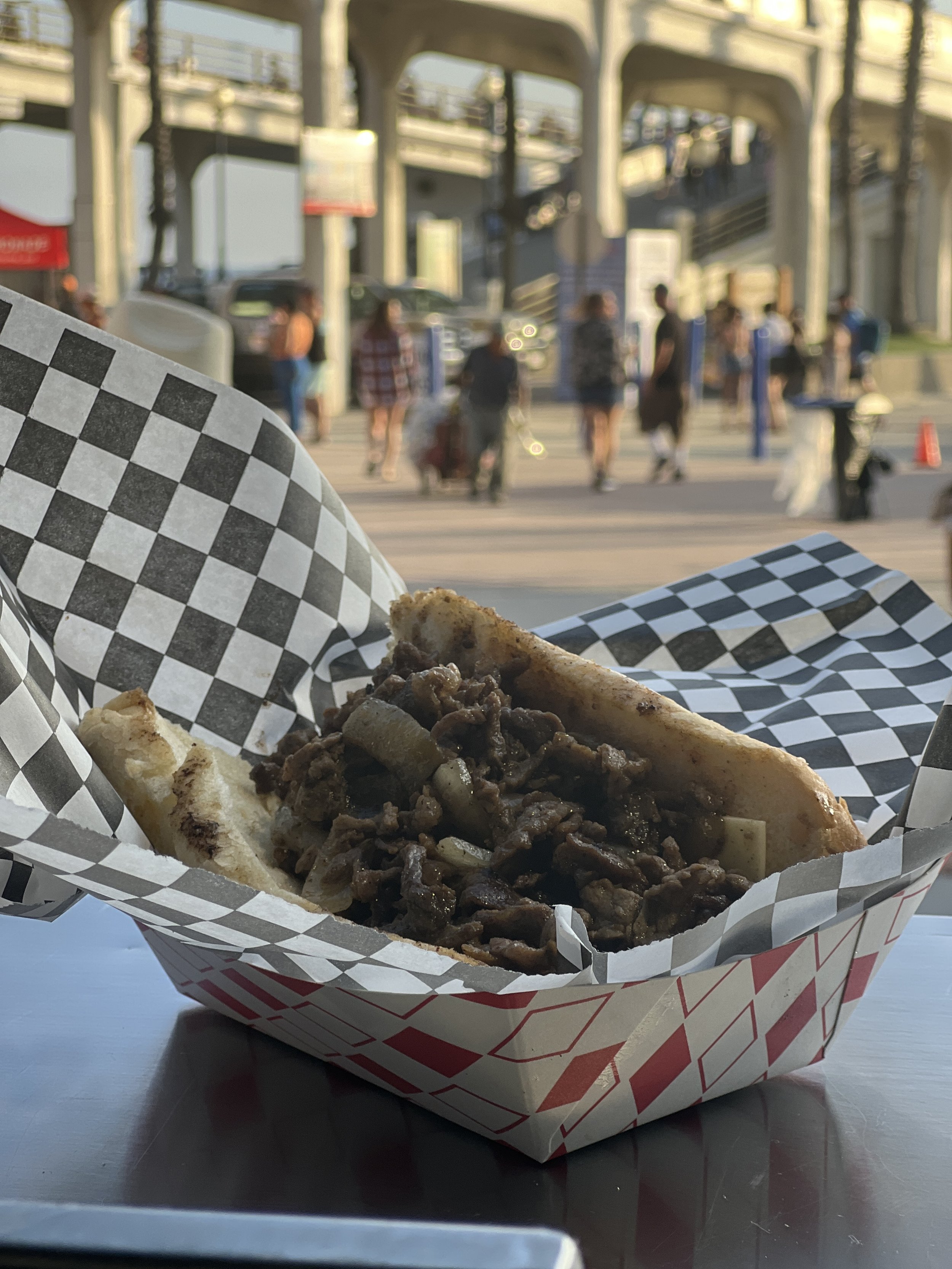 A gyro sandwich filled with cooked beef and onions in a paper tray, set on a table with a city street and bridge in the background.
