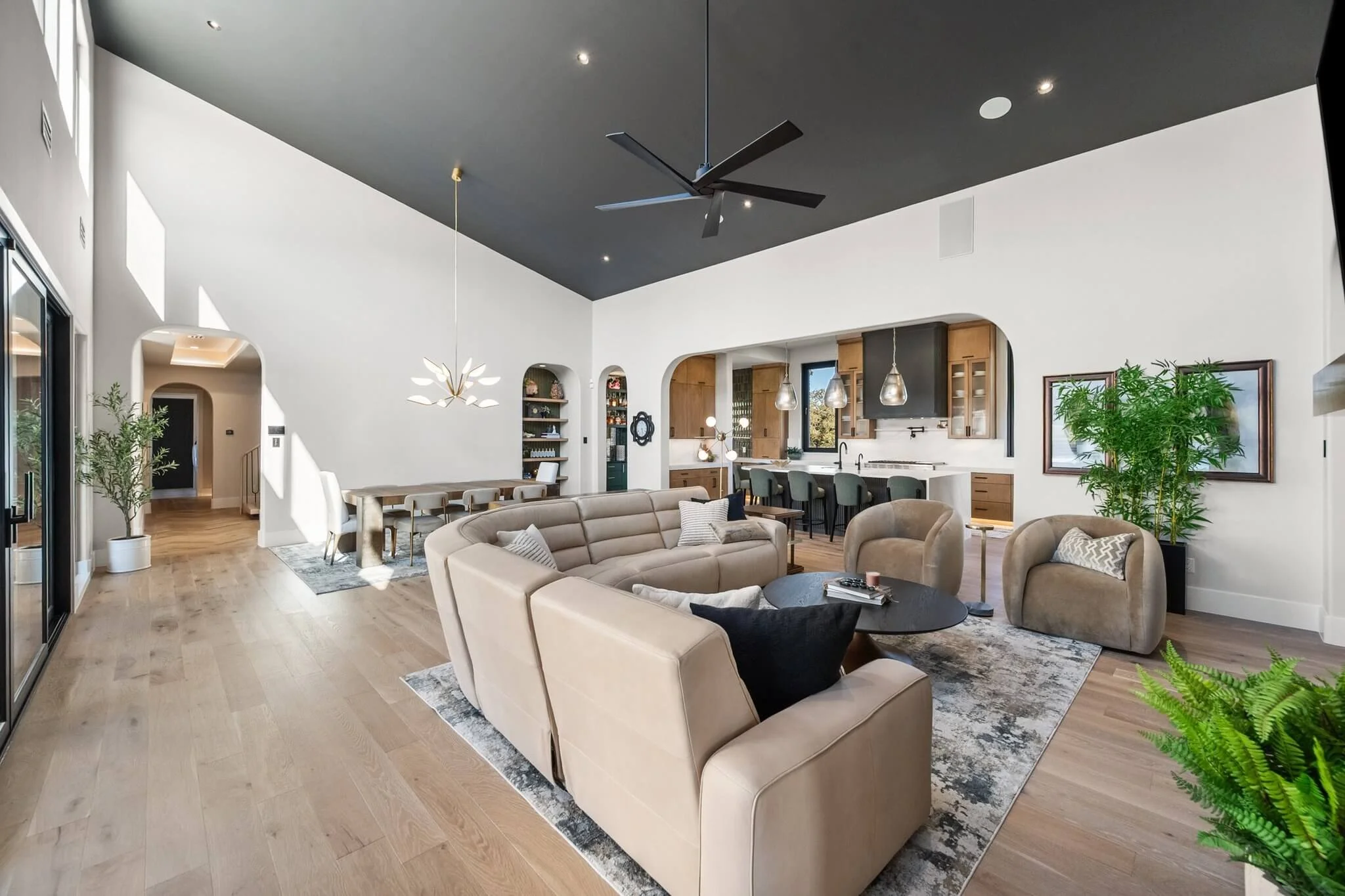 Open-concept living room and kitchen with high ceiling, beige sectional sofa, armchairs, black coffee table, light wood flooring, and modern decor, including green indoor plants.