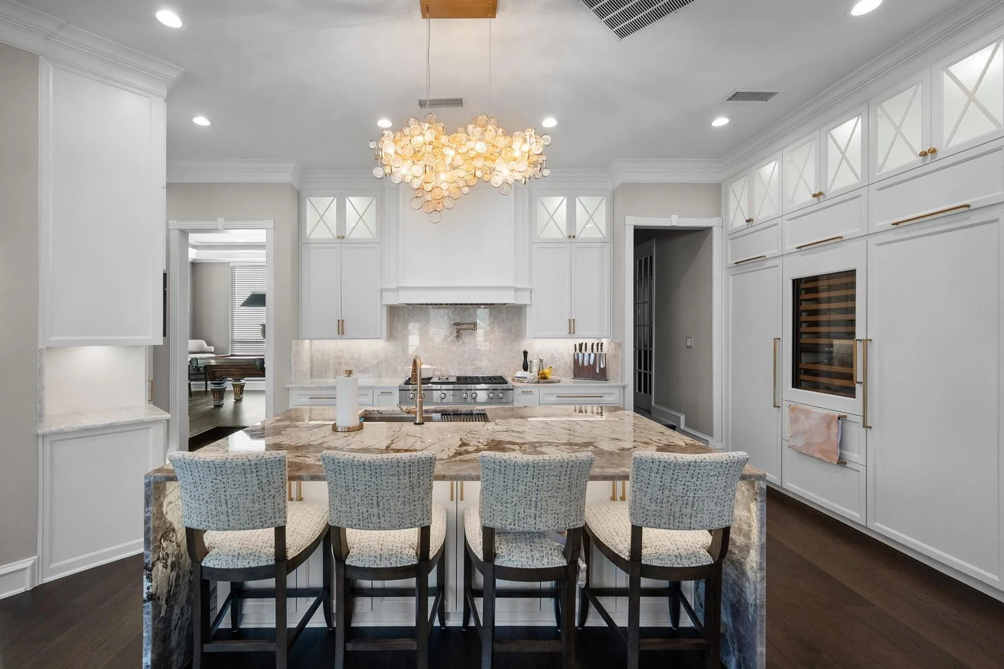 Modern kitchen with white cabinetry, marble island with seating, stove, and a wine fridge. There is a contemporary chandelier hanging from the ceiling and hardwood floors in Mansfield, TX.