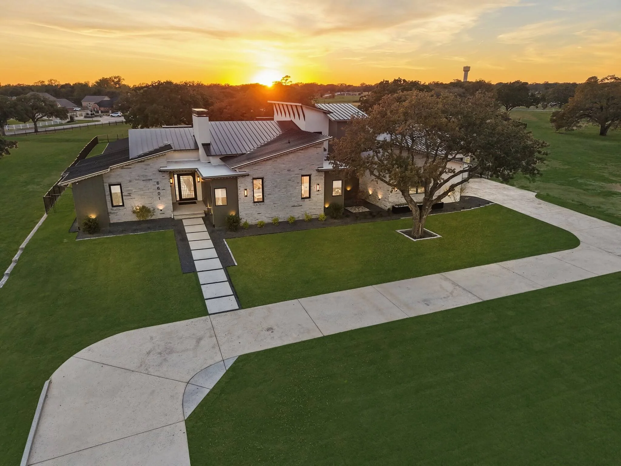 A modern house with a stone exterior and a metal roof, surrounded by a well-maintained lawn, with a large tree in the front yard and a paved driveway curving around it. The sun is setting in the background, casting a warm glow over the scene.