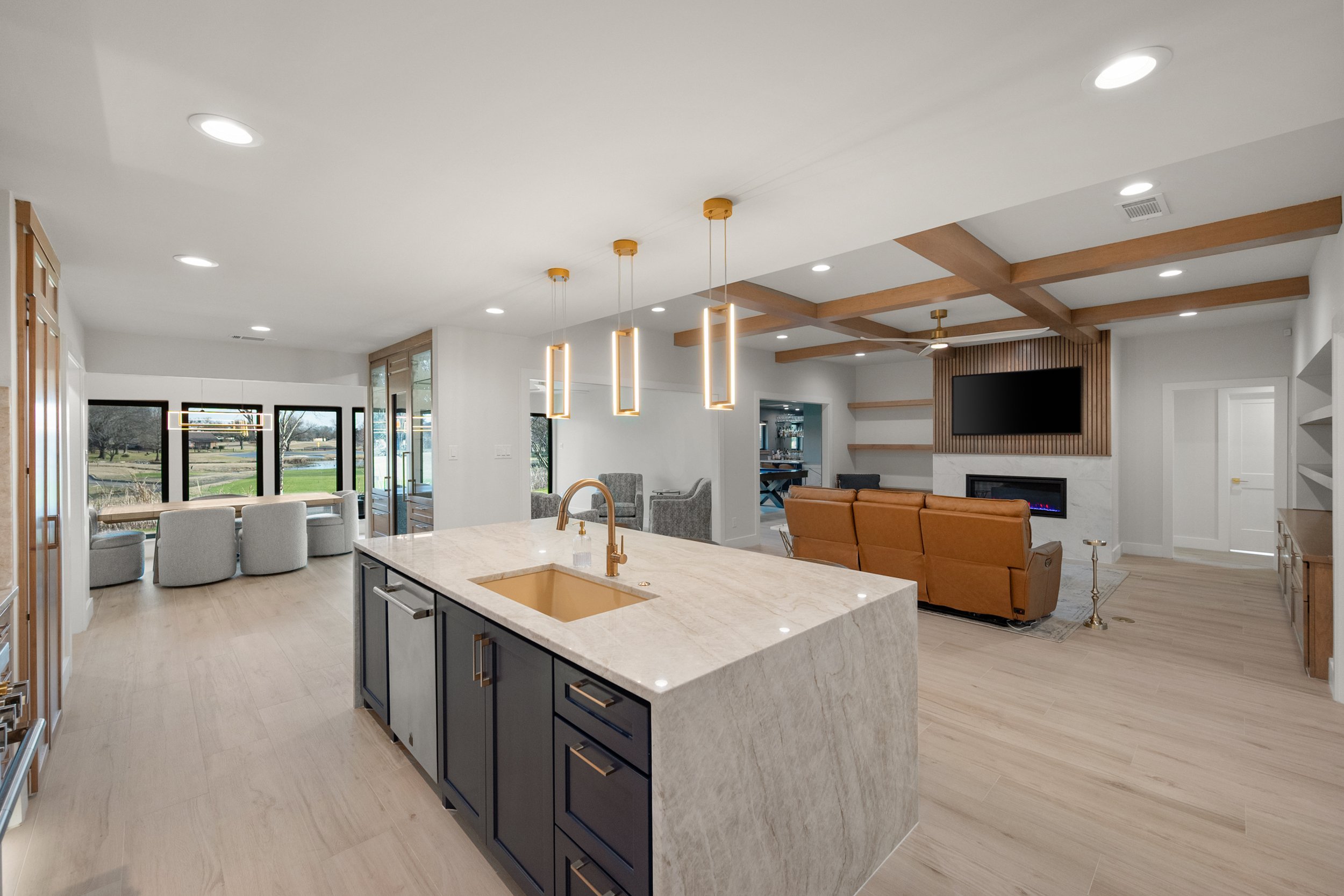 Open-concept living space with kitchen island, seating area, and fireplace with wall-mounted TV, modern furniture, and large windows showing outdoor scenery.