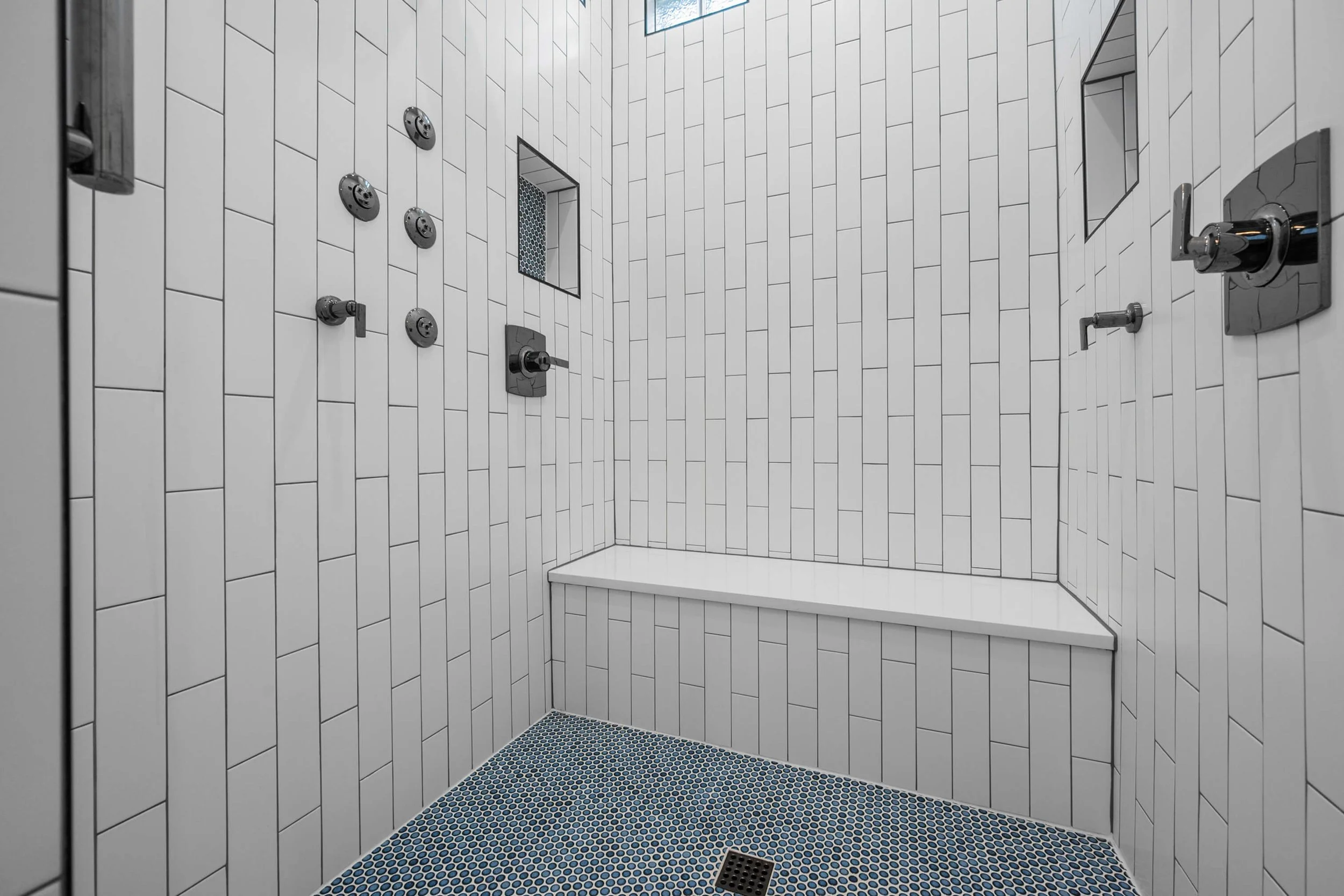 Interior of a modern shower with white rectangular tiles, blue circular mosaic floor tiles, multiple showerheads, a built-in bench, and a small niche for toiletries in Mansfield, TX.