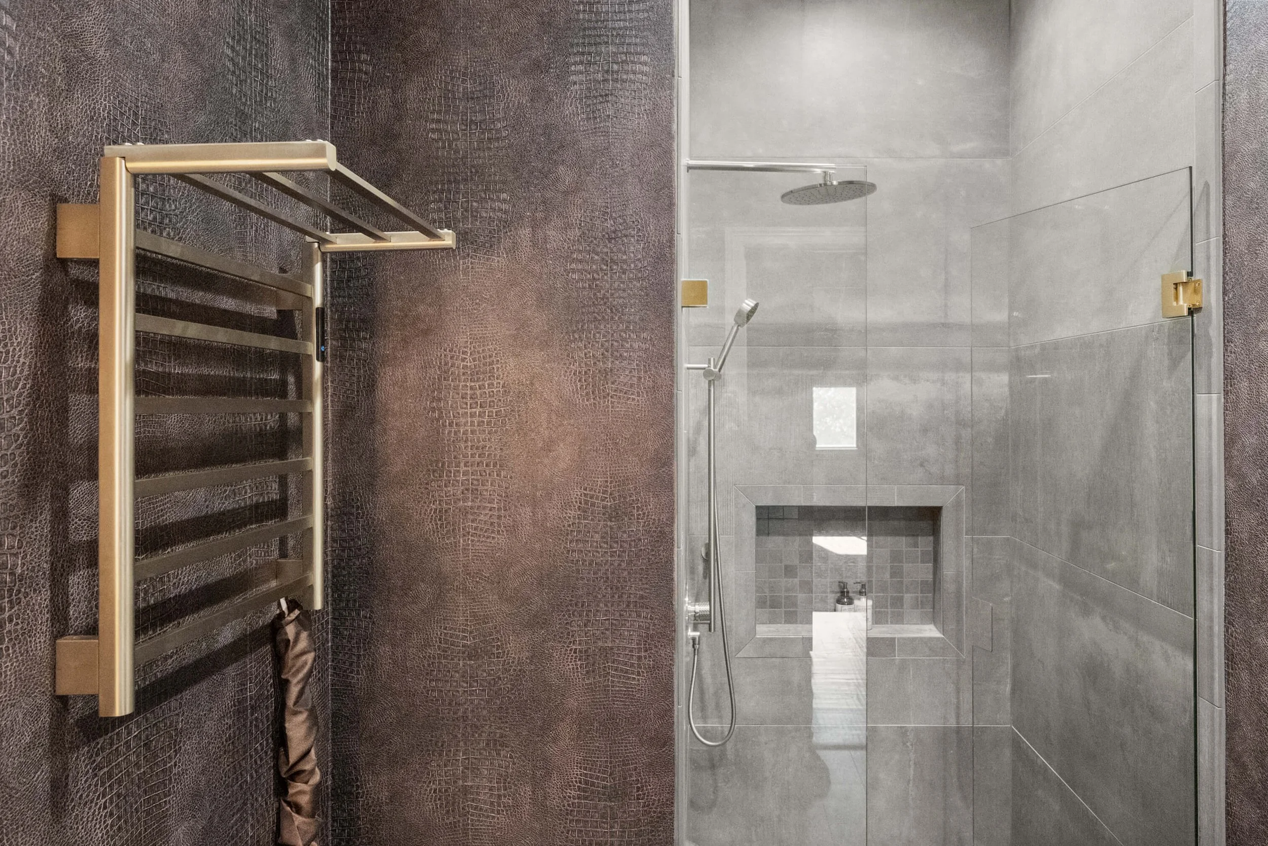 Modern walk-in shower with a glass door, gray tiled walls, built-in niche, and a rain showerhead, with a bronze heated towel rack on a textured dark wall.