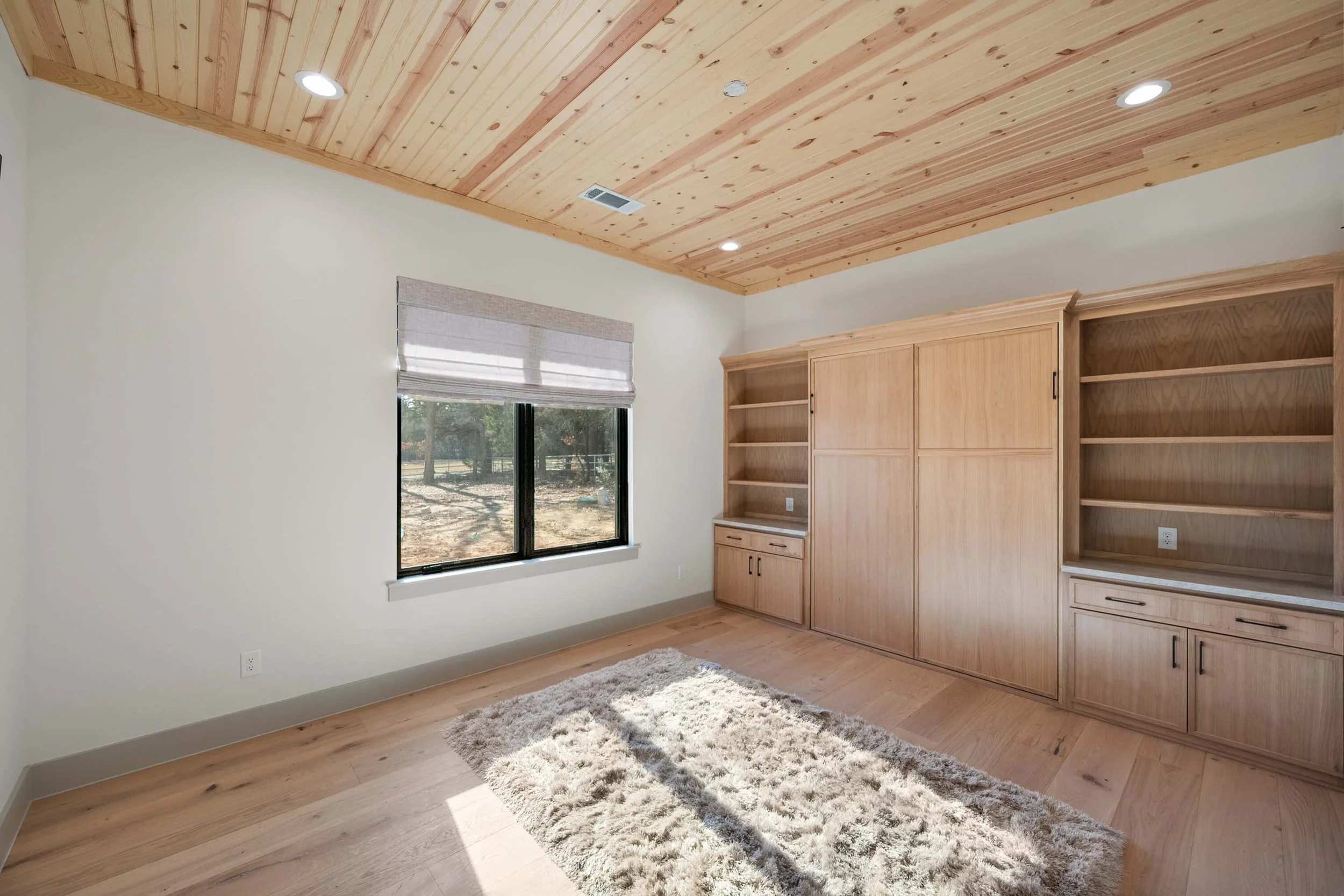 Empty room with beige walls, wooden ceiling with recessed lighting, a window with blinds, a built-in wooden bookshelf and cabinet, and a plush rug on the light wood floor.