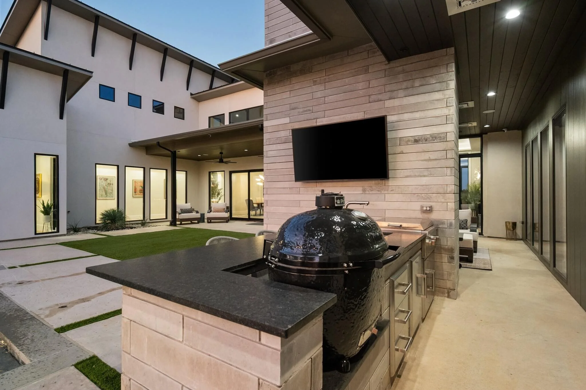 Outdoor patio area of a modern house with a built-in grill, a TV mounted on a brick wall, and view into an indoor living space through sliding glass doors.