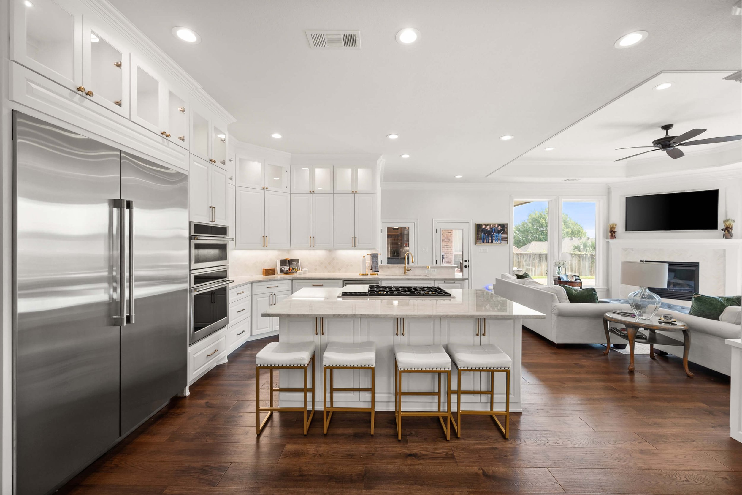 Bright modern open-concept kitchen and living room with white cabinets, marble countertops, stainless steel appliances, a kitchen island with white stools, dark wood flooring, a white sofa set, a wall-mounted TV, and large windows letting in natural 