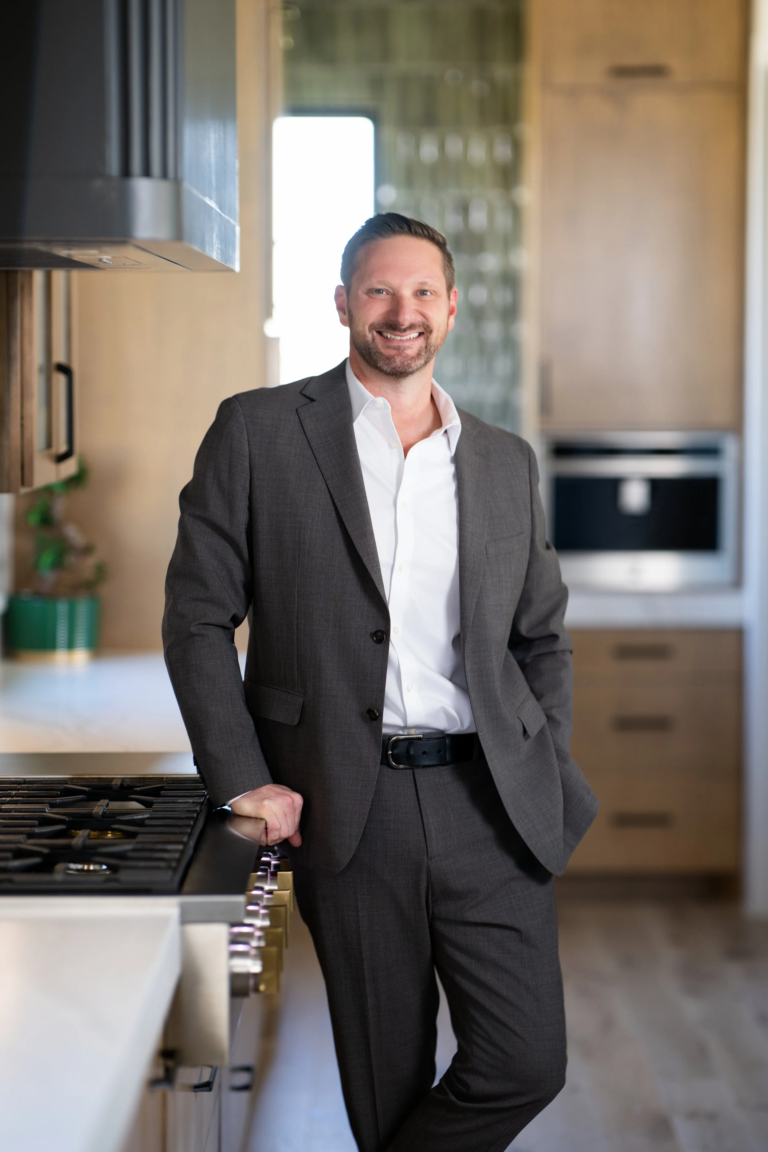 A smiling man in a gray suit and white shirt stands in a modern kitchen, leaning against a stove with one hand on the countertop.