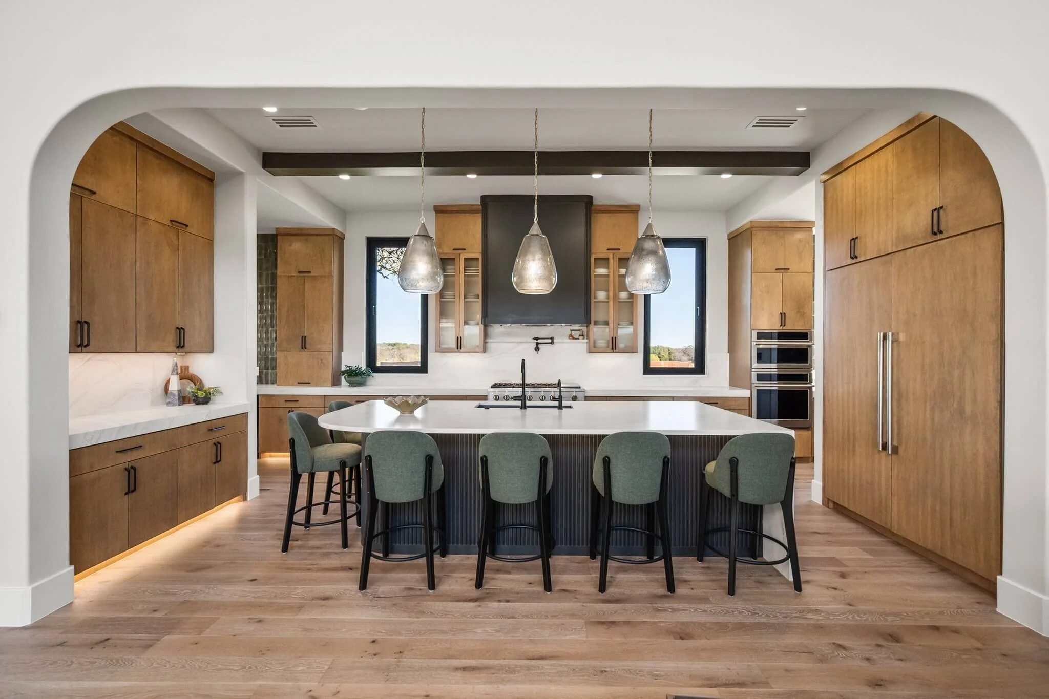 Modern kitchen with wooden cabinets, a large white island with seating, three pendant lights above, black-framed windows, and built-in appliances.