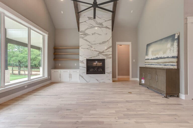 Empty living room with large window, modern fireplace with white marble surround, built-in shelves, and neutral-colored furniture.