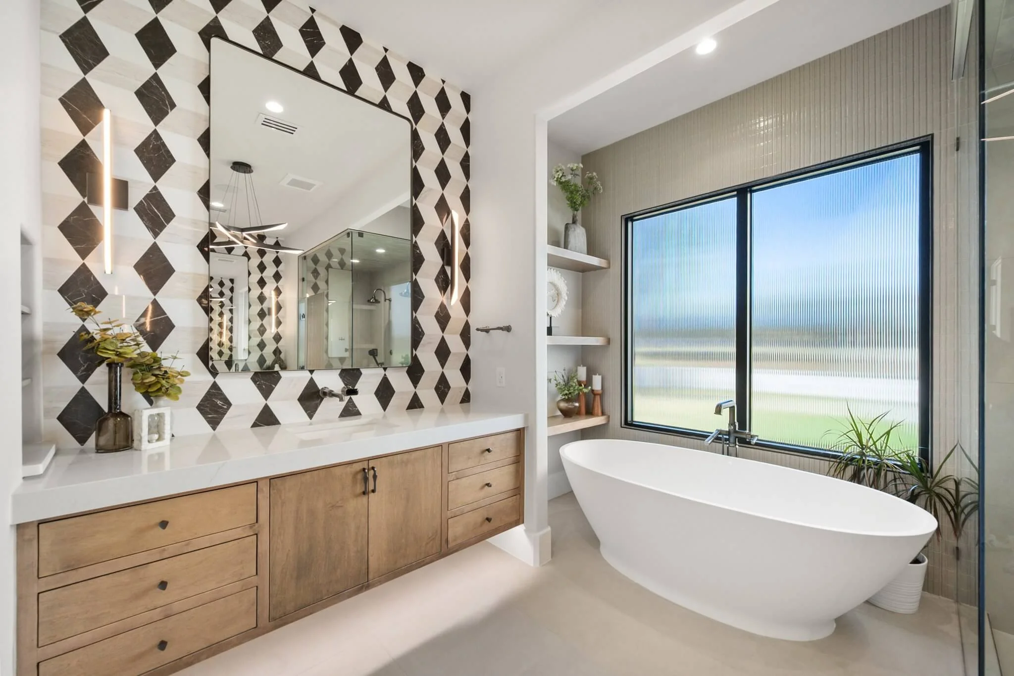Modern bathroom with a freestanding bathtub in front of a large window, a wooden vanity with a white countertop and a geometric black and white tiled wall, and built-in shelves with decorative items.