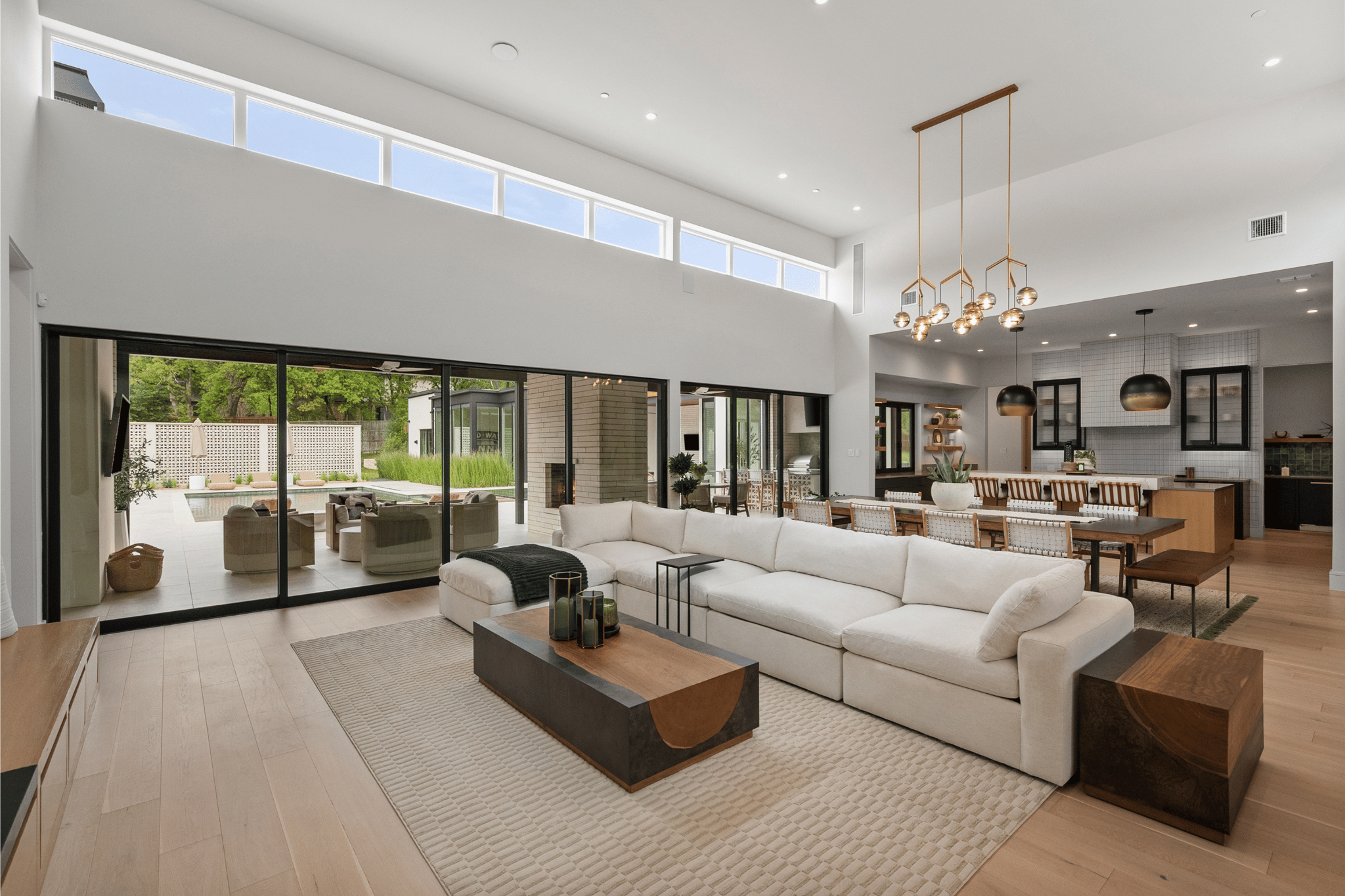 Modern open-concept living room with large white sectional sofa, wooden coffee table, and sliding glass doors leading to outdoor patio with seating and greenery.