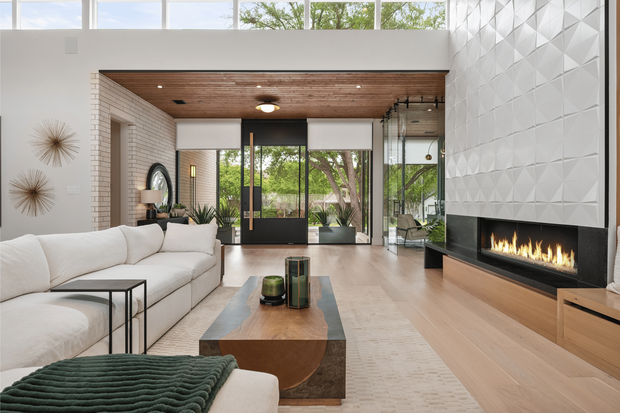 Modern living room with white sofa, wooden coffee table, gas fireplace, plants, and large glass doors leading outside to a green yard.