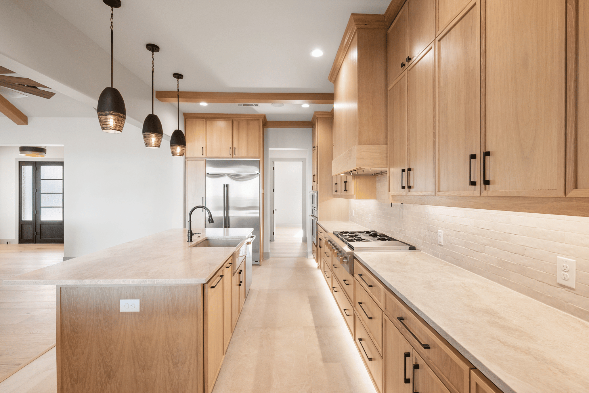 Modern kitchen with light wood cabinets, beige countertops, stainless steel appliances, and black pendant lights.