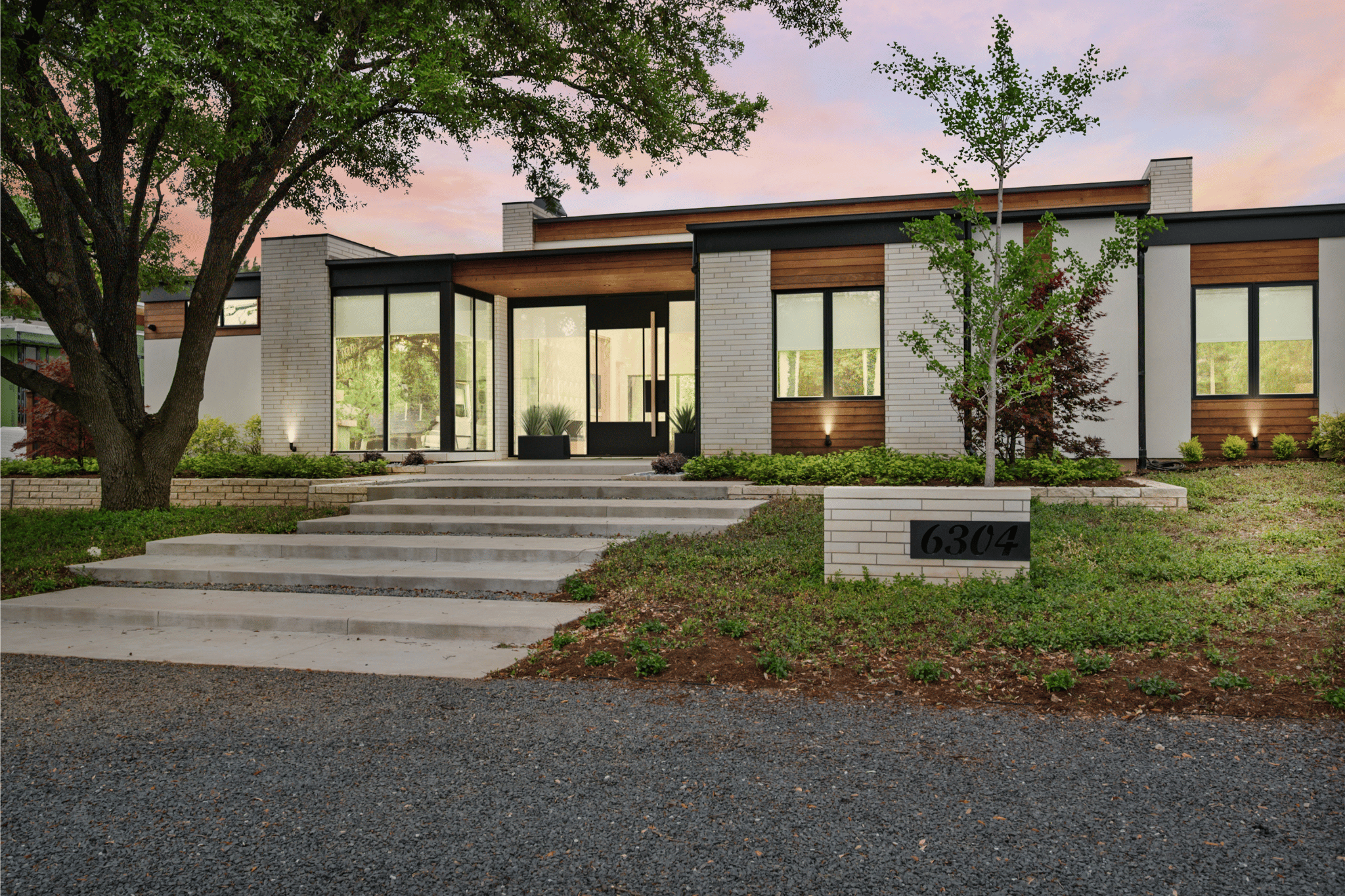 A modern house with white brick walls, large glass windows, and wooden accents, surrounded by landscaped green grass and trees, with a concrete pathway and steps leading to the front door, and house number 6304 on a corner brick sign.