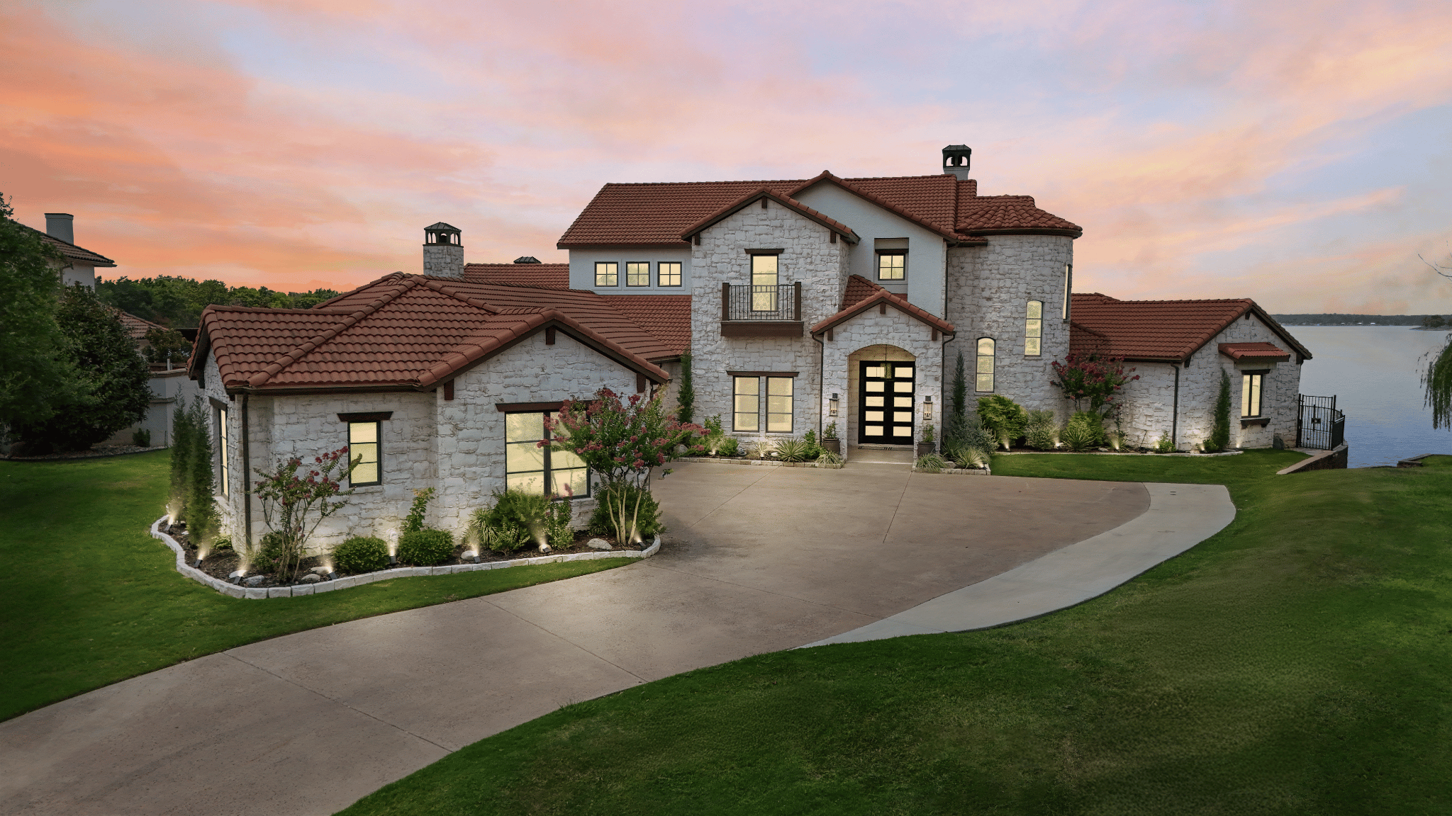 A large, luxurious house with stone walls and red tile roof, situated near water, during sunset with a colorful sky. It has landscaped front yard, driveway, and outdoor lighting.