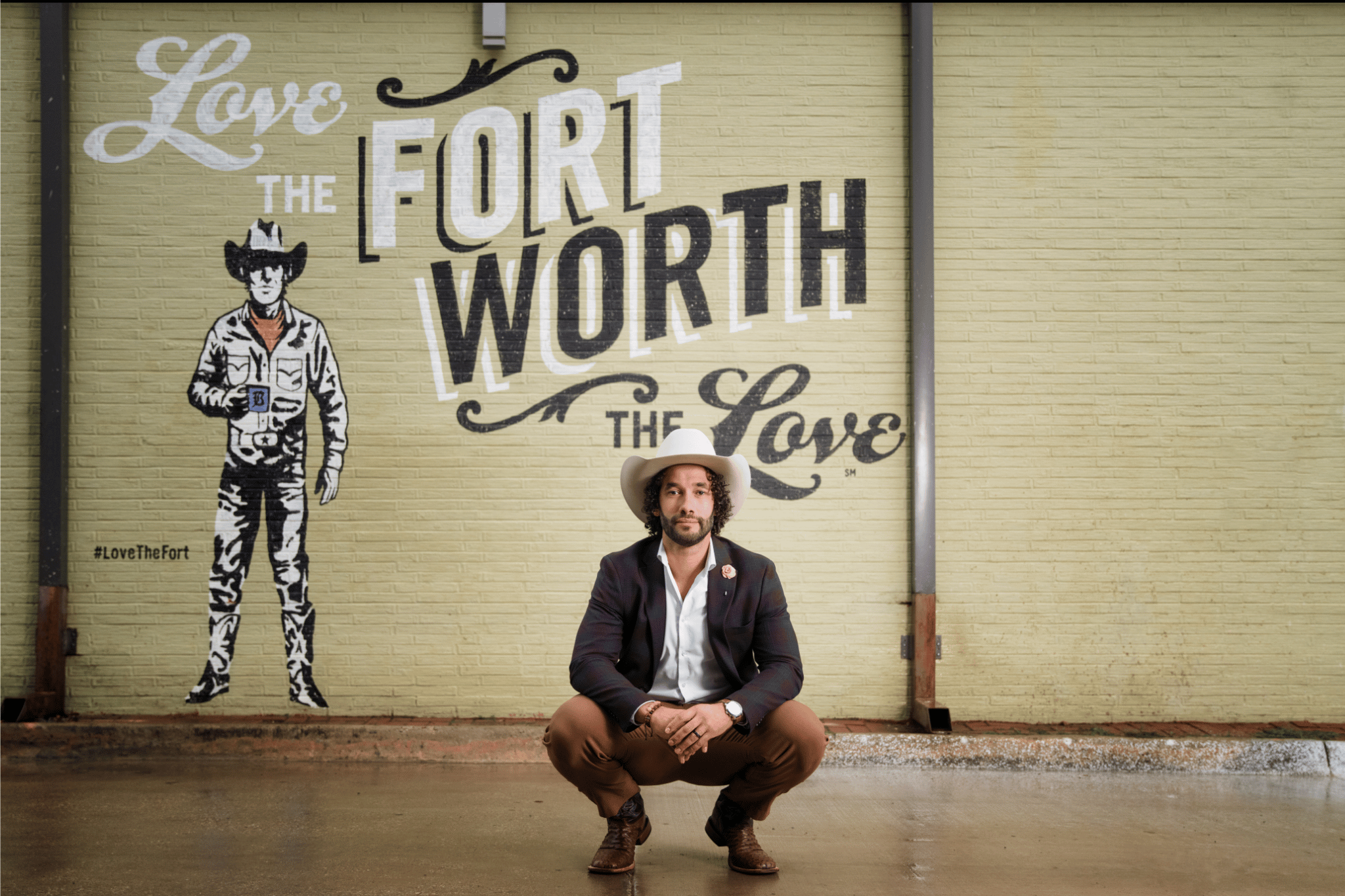 A man wearing a white cowboy hat, white shirt, dark blazer, brown pants, and brown boots is squatting in front of a yellow brick wall with a mural. The mural reads 'Love the Fort, The Fort is the Love' and features an illustration of a person in cowboy attire holding a mug, with the hashtag #LoveTheFort.
