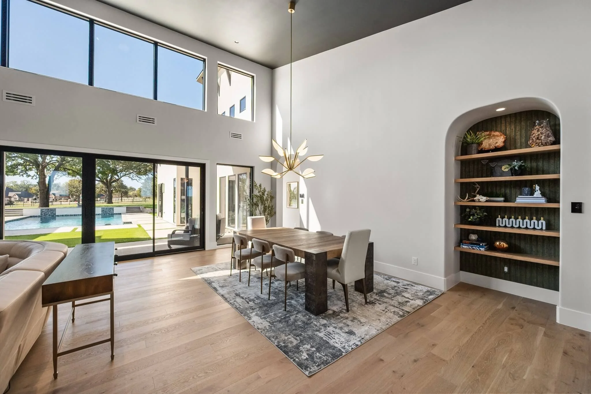 Modern dining room with large windows, wooden floor, dining table with six chairs, built-in shelves with decorations, and a sliding glass door leading to a backyard with a pool and outdoor seating.