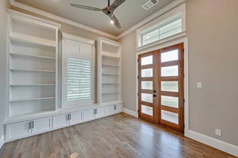 Empty room with built-in white bookshelves, a window with shutters, a glass-paneled wooden door, hardwood flooring, and a ceiling fan.