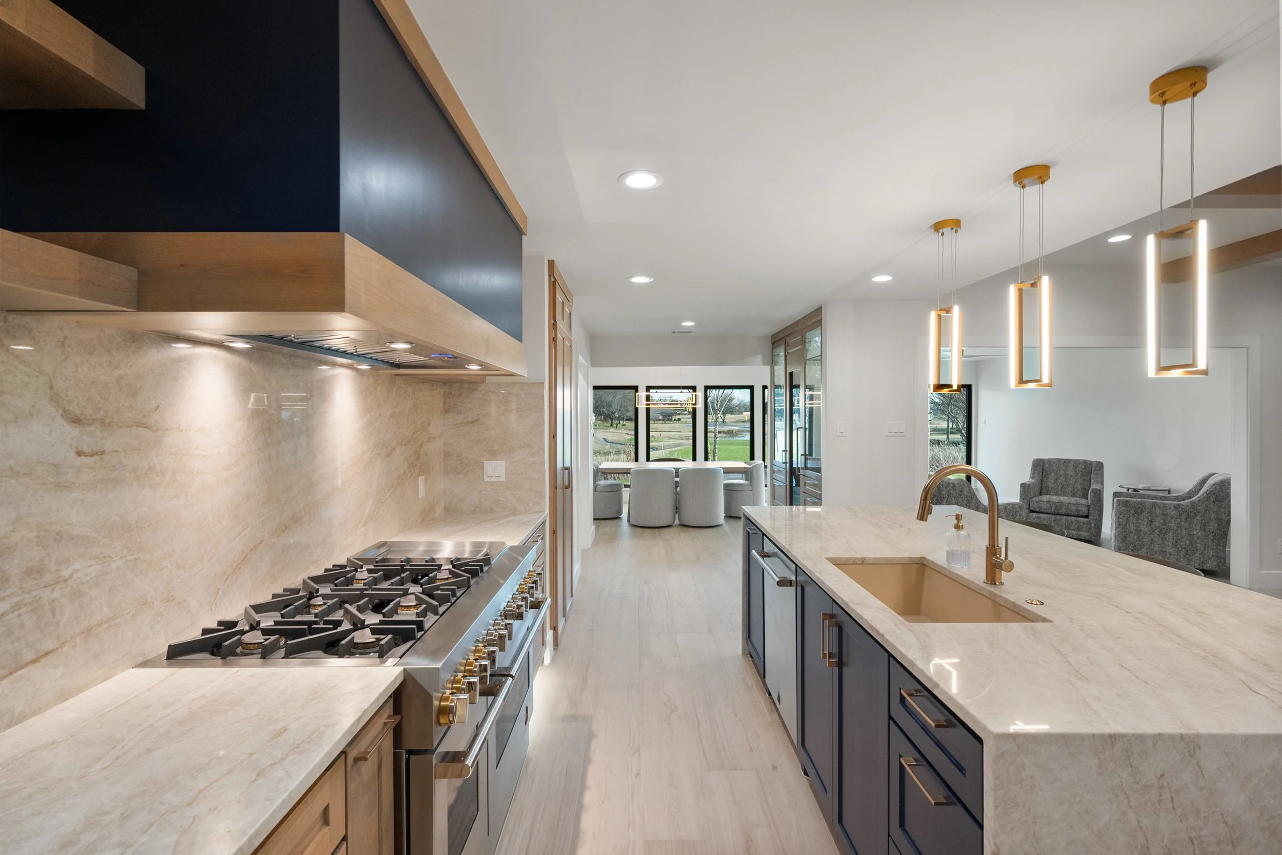 Modern open-concept kitchen with marble countertops, gold fixtures, and pendant lighting, flowing into a living room with gray armchairs and a view of a yard through large windows built by Lakeland Custom Homes.