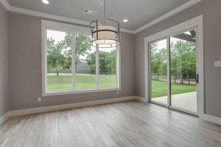 Empty room with large window and sliding glass door, light wood flooring, gray walls, and a modern ceiling light fixture, overlooking a backyard with trees and a fence.