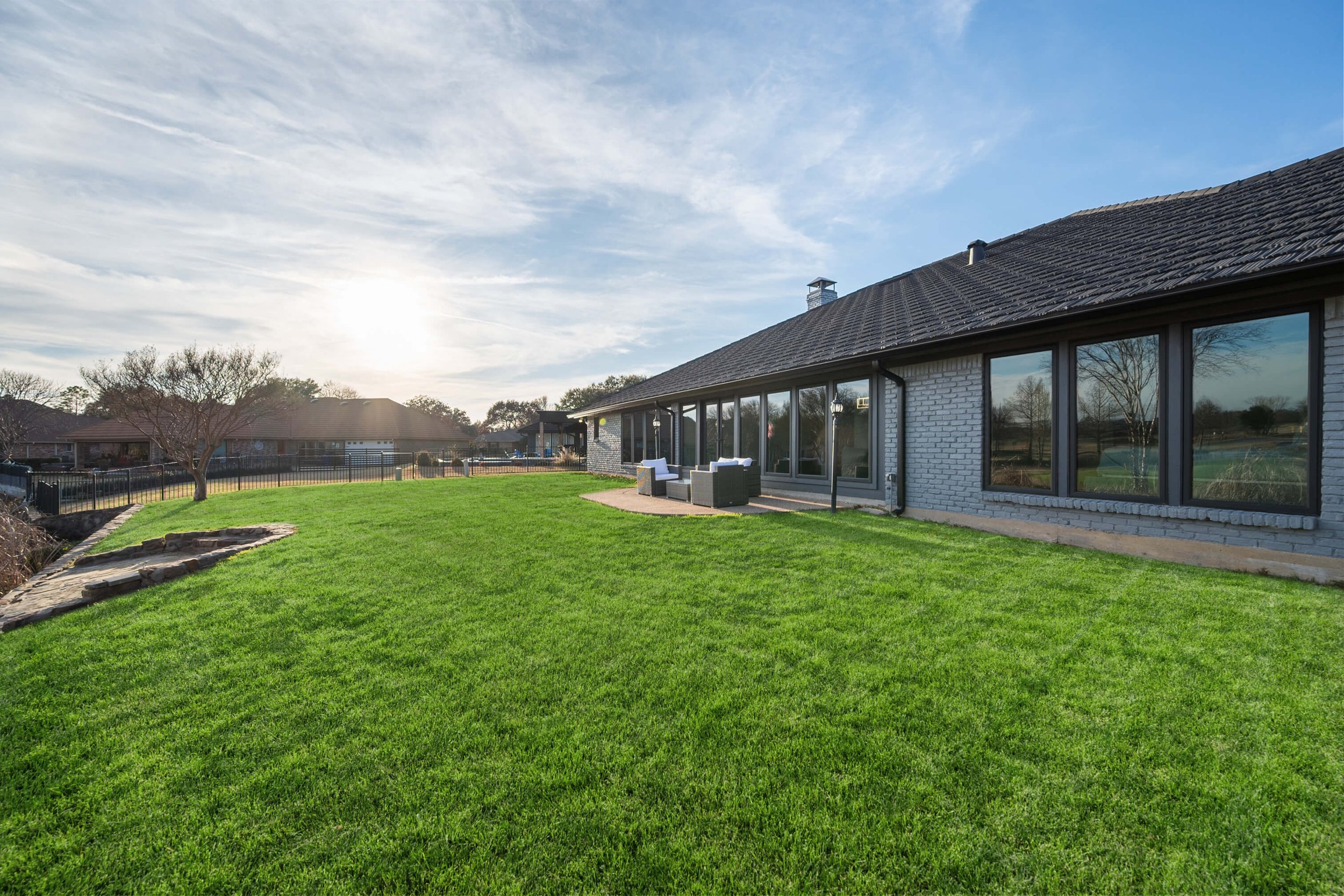 Backyard of a modern house with large windows, green lawn, patio furniture, and a few trees under a sunny sky.