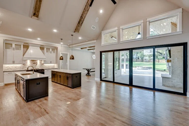 Empty modern open-concept kitchen and living area with hardwood floors, white cabinets, a large island, pendant lighting, and large glass sliding doors leading outside in Mansfield, TX.