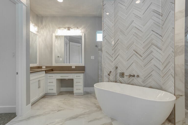 Modern bathroom with a white freestanding bathtub, herringbone tile feature wall, marble flooring, and a vanity with a large mirror.