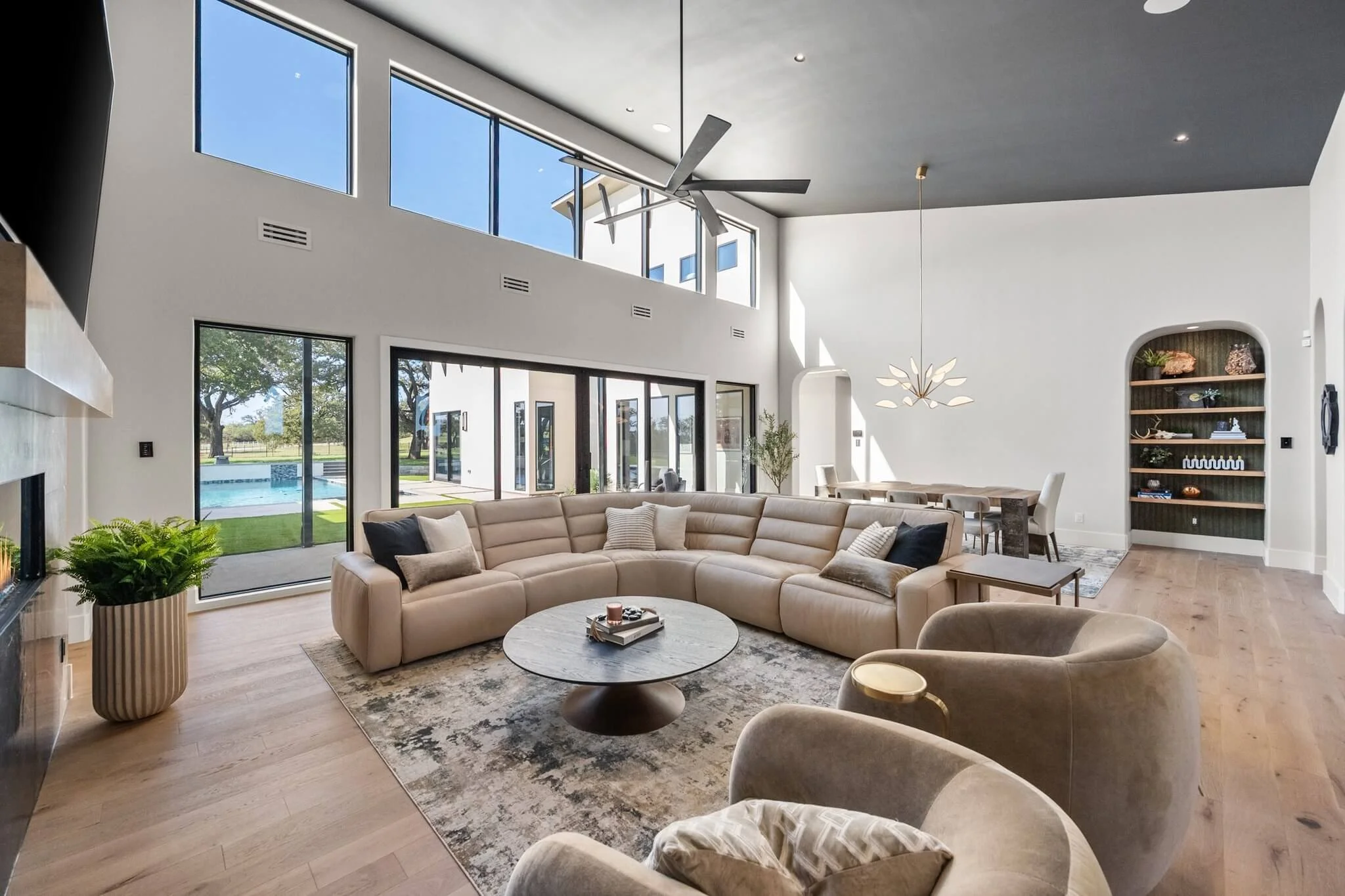 Modern living room with high ceiling, large windows, a sectional sofa, armchairs, a round marble coffee table, a fireplace, and a view of the backyard with a pool.