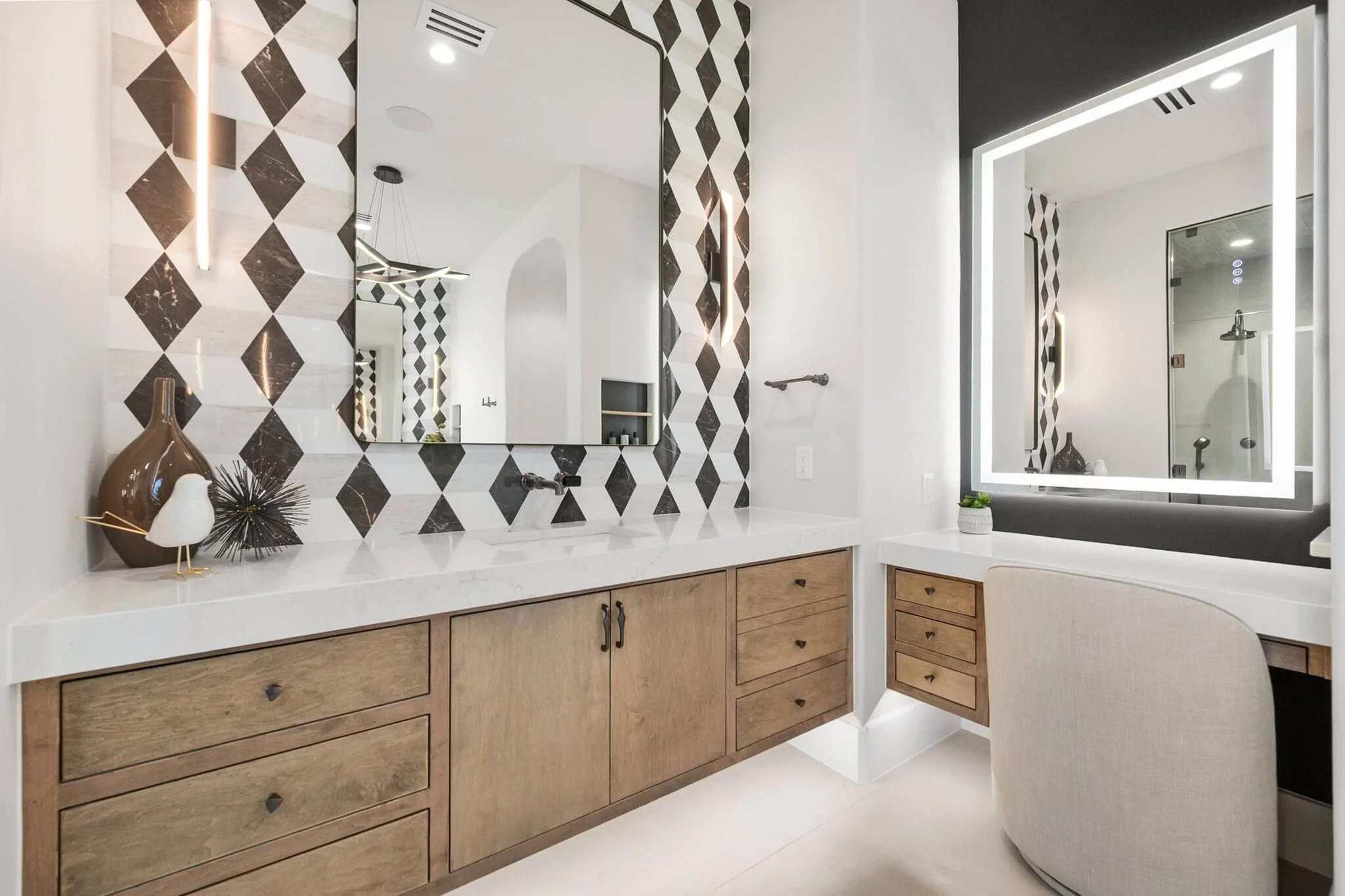 Modern bathroom with black and white diamond-patterned wall tiles, a large illuminated mirror, wooden vanity with white countertop, decorative vases and bird sculpture, and a small built-in desk area with a chair.