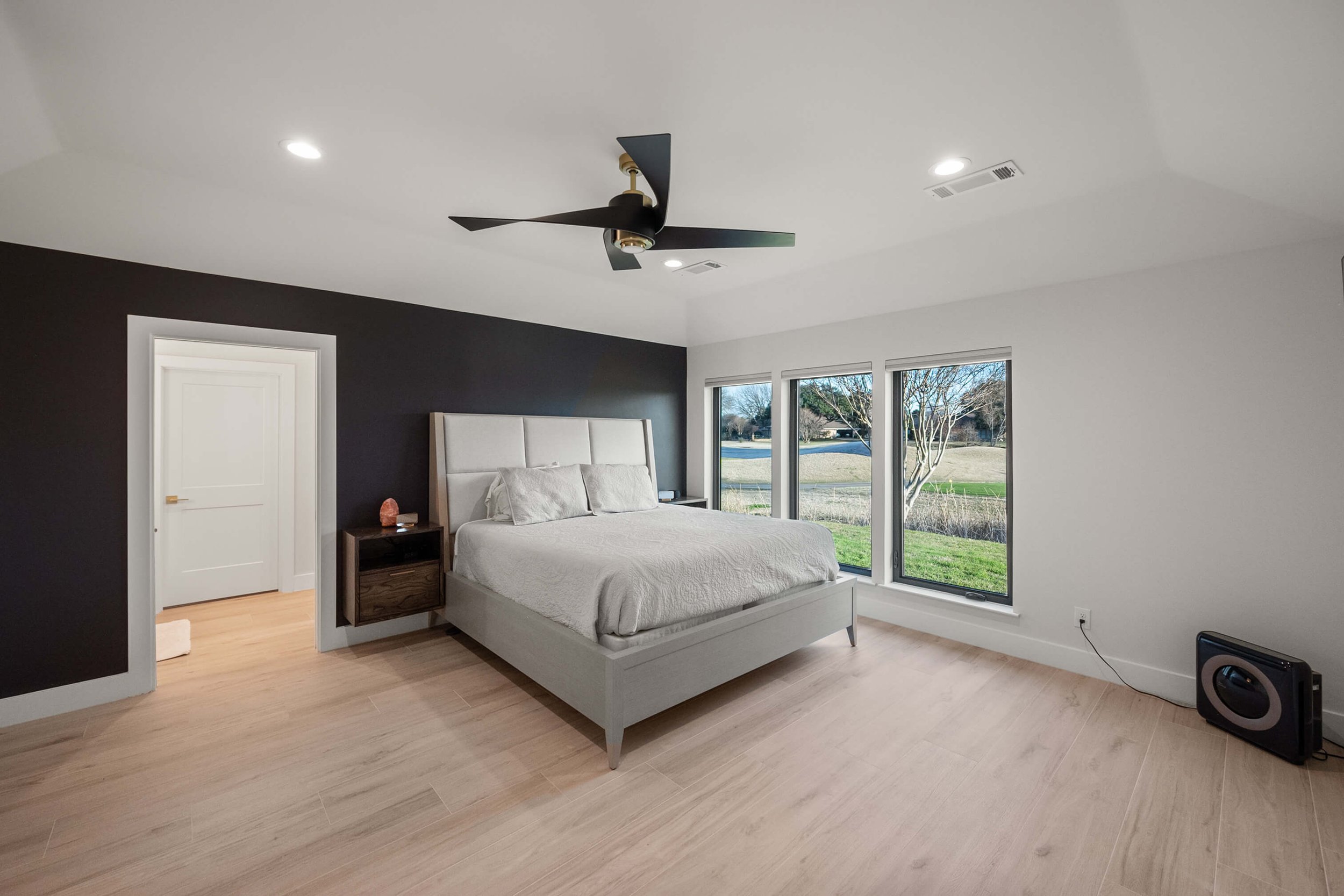 A modern bedroom with a white bed, black wall, wooden nightstand, large windows with a view of a yard, ceiling fan, and light wood flooring.