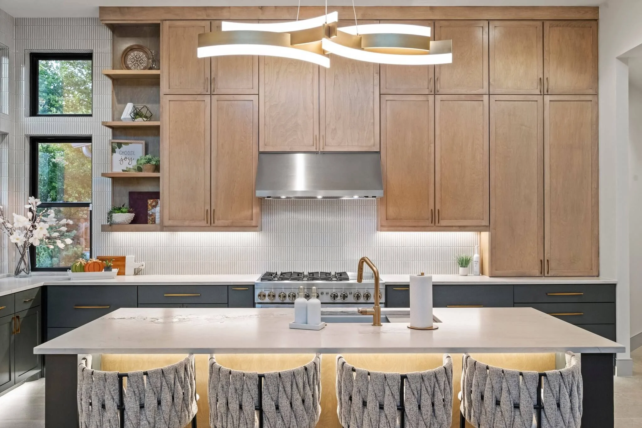 Modern kitchen with light wood upper cabinets, dark lower cabinets, white countertop, a central island with a white surface, and a stainless steel range hood. Decorative open shelving on the left wall, and a contemporary gold and white chandelier ove