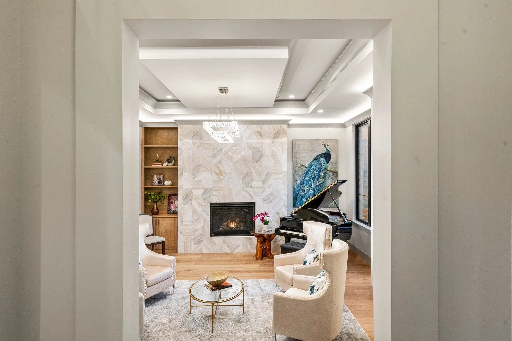 Living room with beige armchairs, a glass coffee table, a black grand piano, a peacock painting, a fireplace, and built-in wooden shelves with decorative items.
