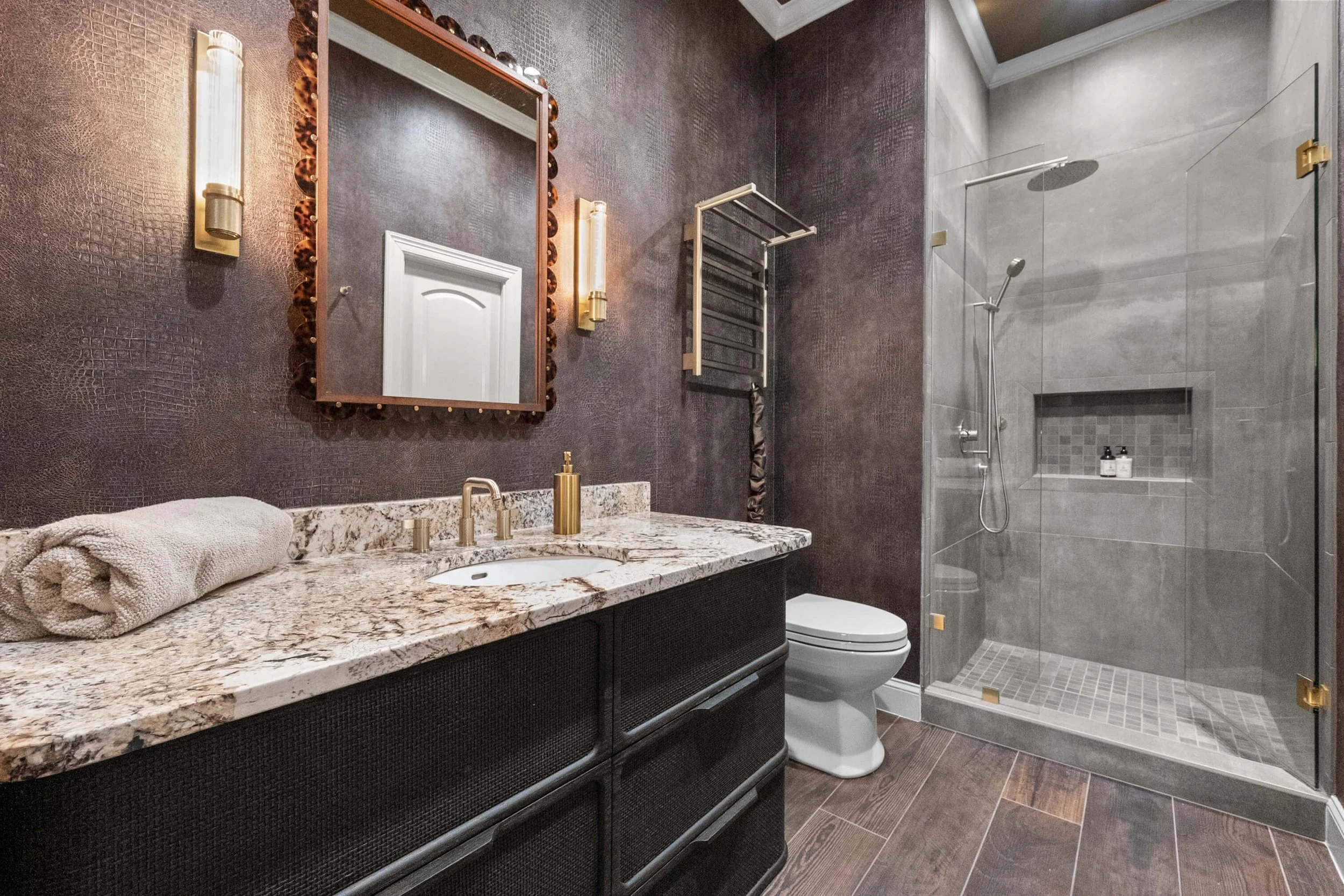 Modern bathroom with a granite countertop vanity, a mirror with a decorative frame, two wall-mounted lights, a towel rolled on the counter, a toilet, and a glass-enclosed shower with gray tiles and a wall-mounted showerhead.