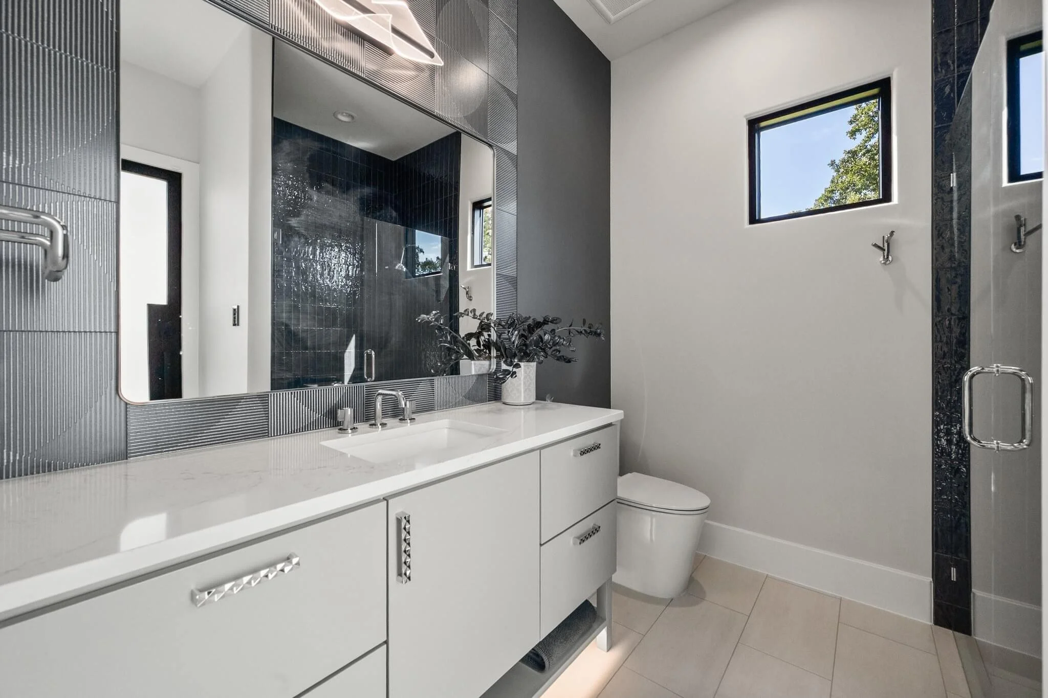 Modern bathroom with white vanity, large mirror, black tiled shower, and two small windows showing blue sky and trees.