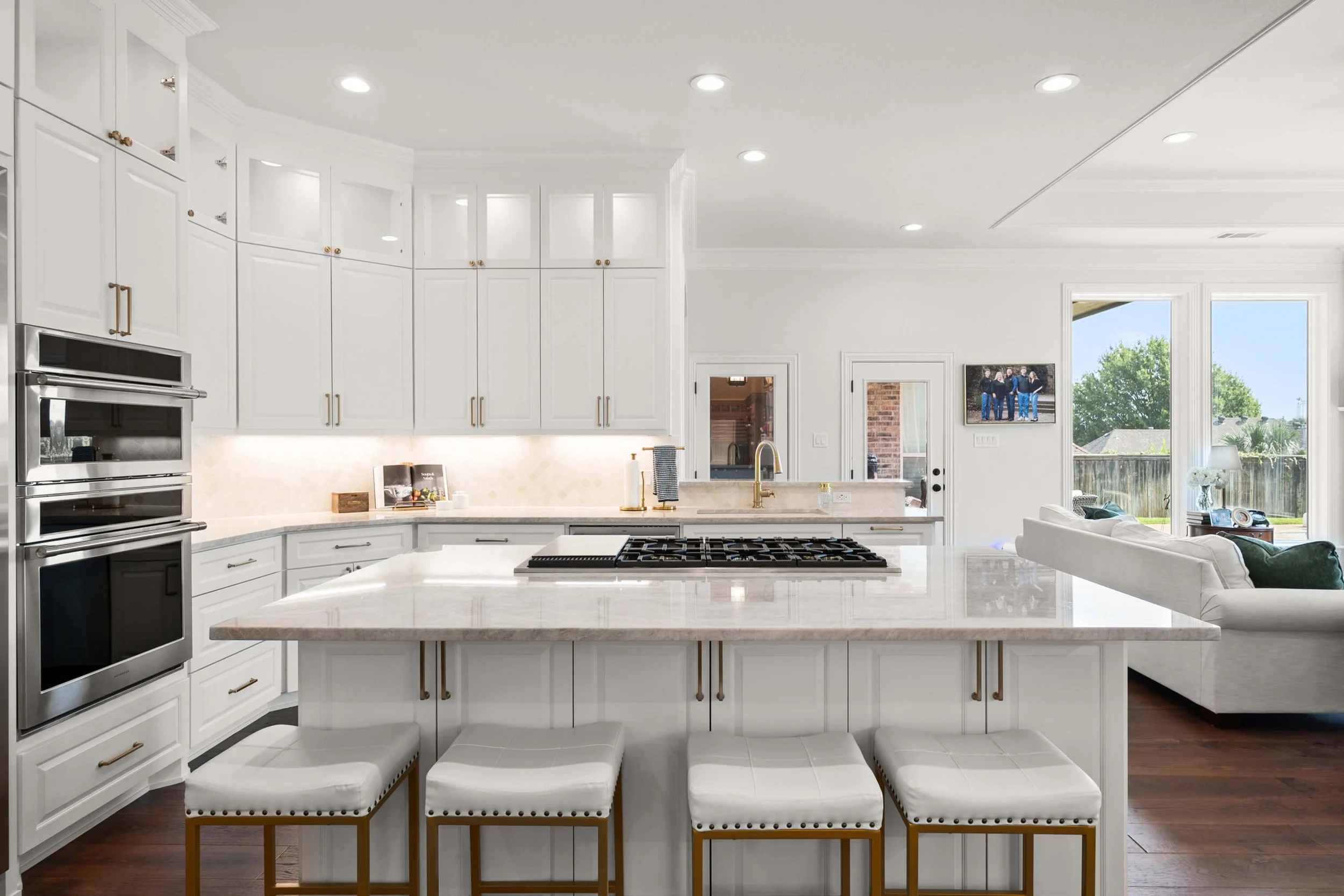 Modern white kitchen with island, four white cushioned bar stools, gold hardware, stainless steel appliances, and open living area with large windows and a backyard view.