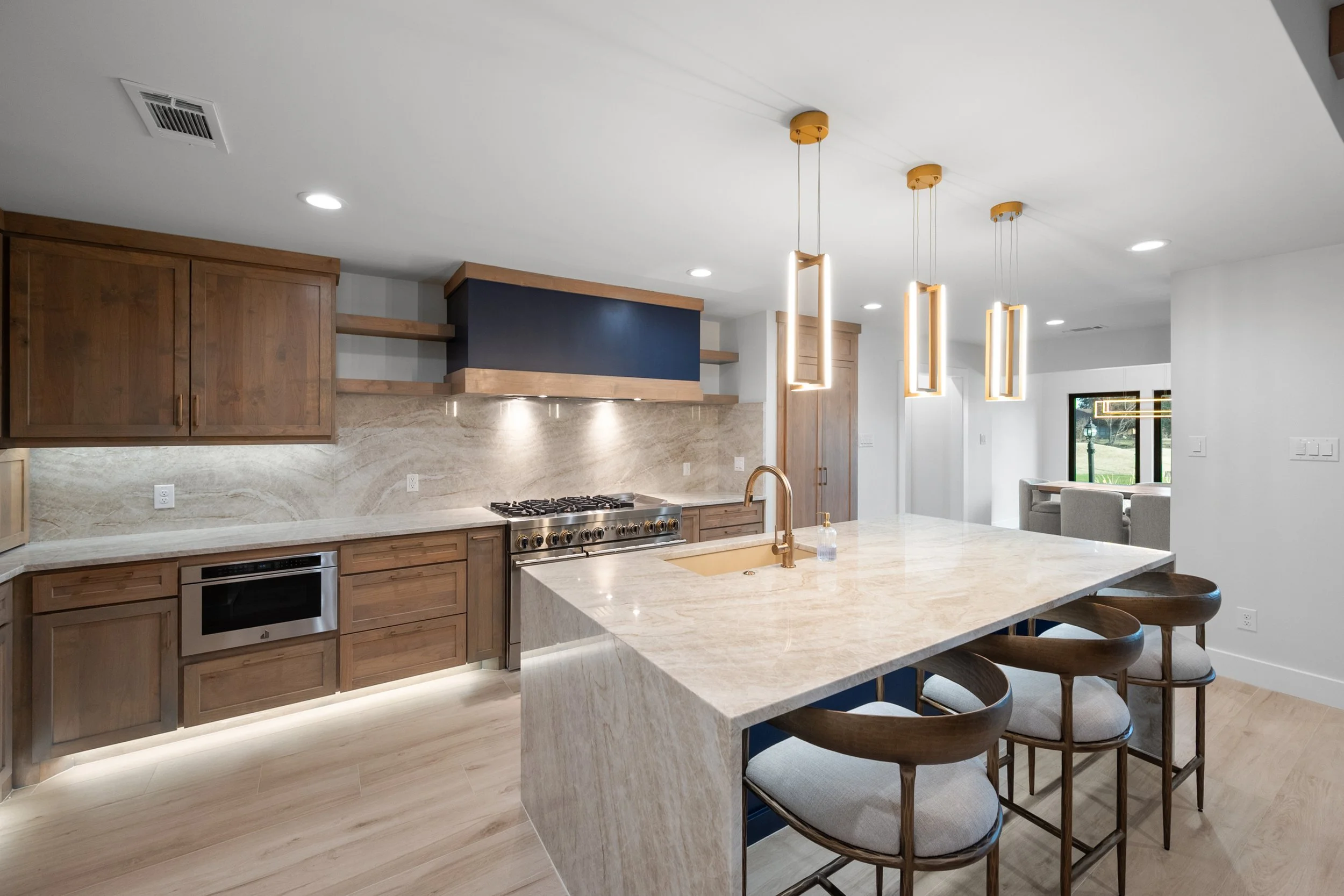 Modern kitchen with wooden cabinets, marble countertops, and a large island with four barstools. Pendant lights hang over the island, and there is a stainless steel oven and microwave. Light-colored hardwood floors and a view of a dining area with a 