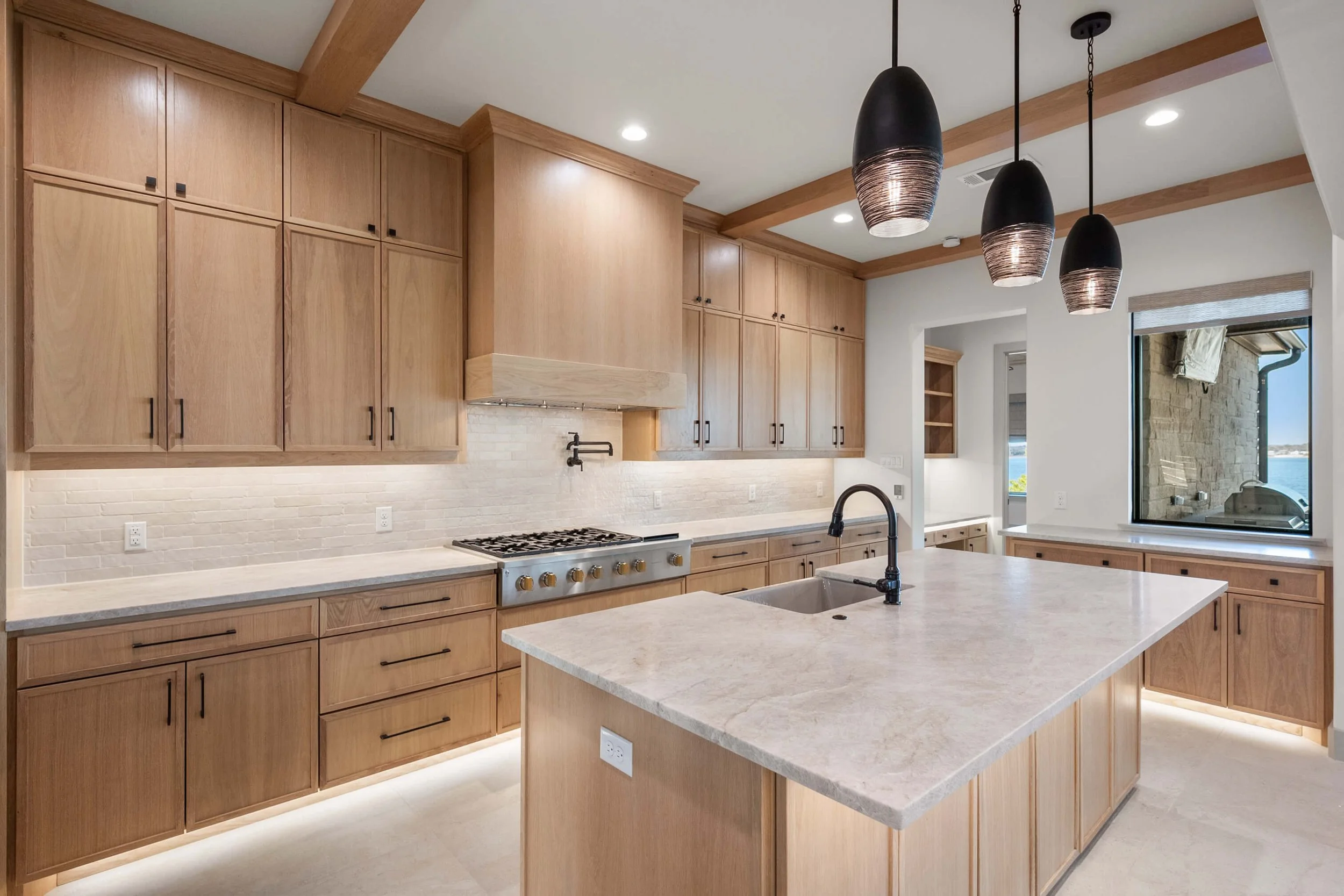 Modern kitchen with light wooden cabinets, a large island with a marble countertop, black pendant lights, a white brick backsplash, a built-in oven, and a window overlooking a patio with water view.