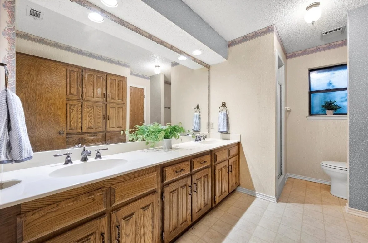Bathroom with double sink vanity, large mirror, wooden cabinets, towel holders, a potted plant, a window, and a toilet.