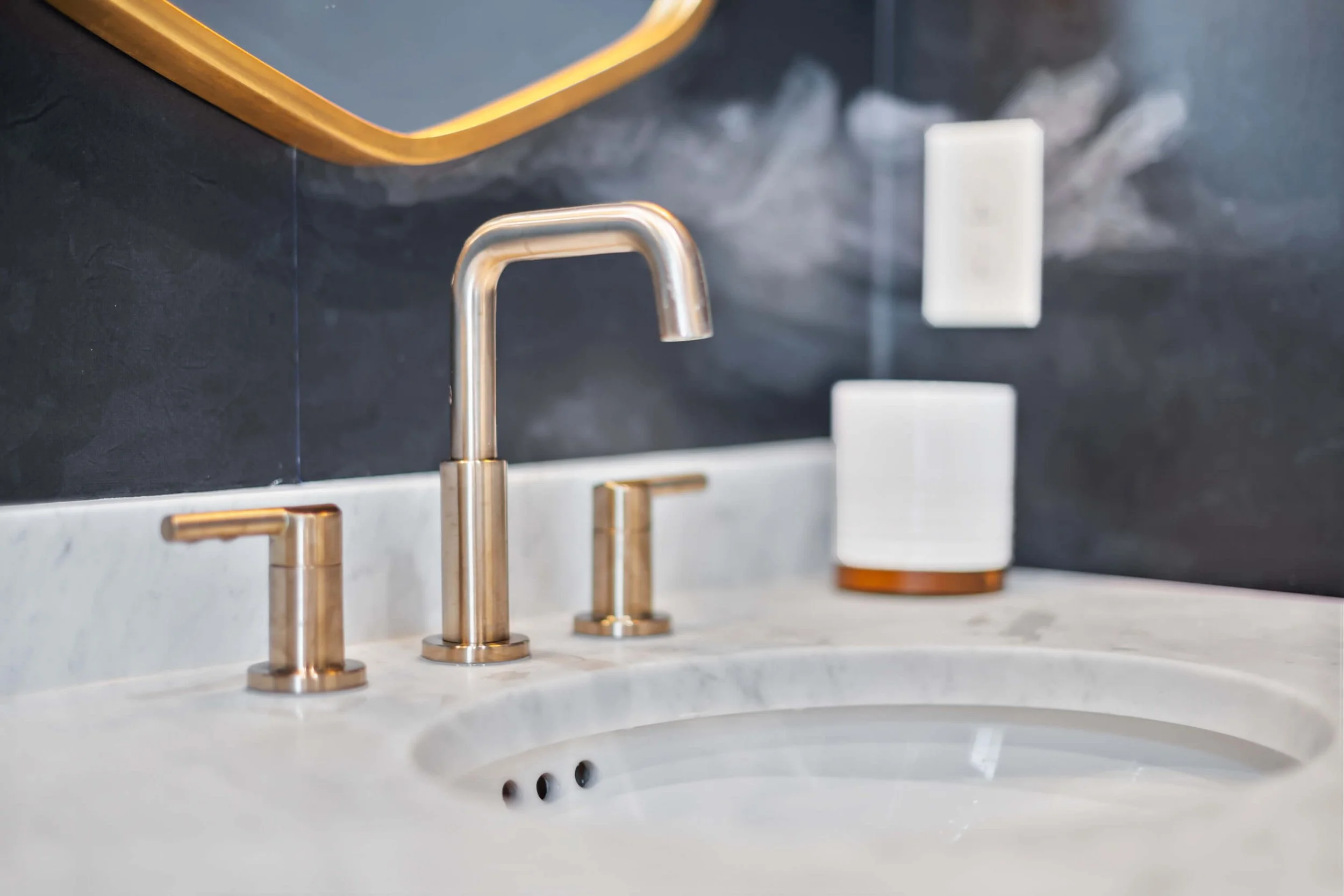 A bathroom sink with a gold faucet and handles, a soap dispenser, and a mirror mounted on dark wall tiles built by Lakeland Custom Homes..