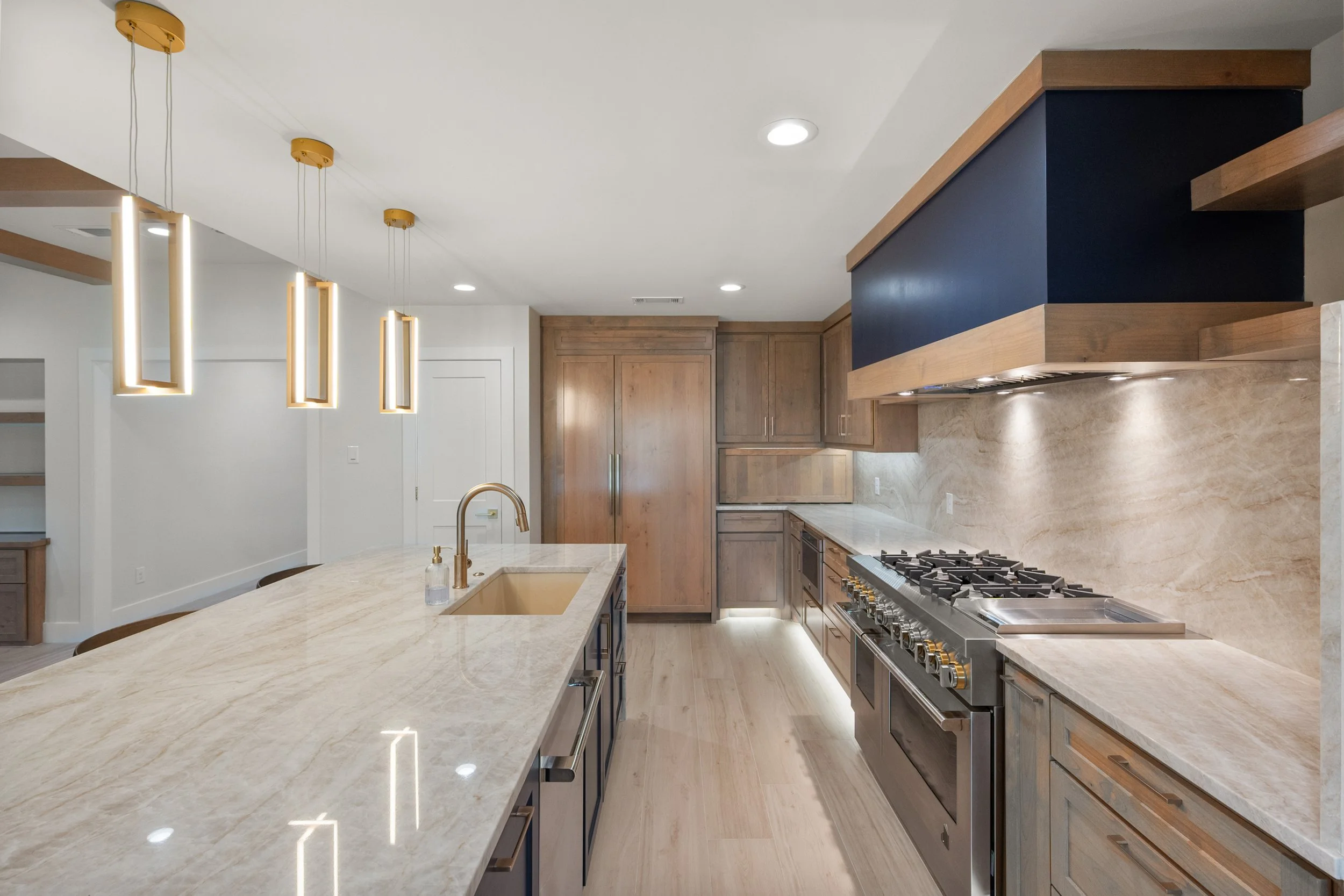 Modern kitchen with beige marble countertops, wooden cabinets, and a stainless steel stove. Features gold fixtures, pendant lights, and a dark blue accent above the stove.