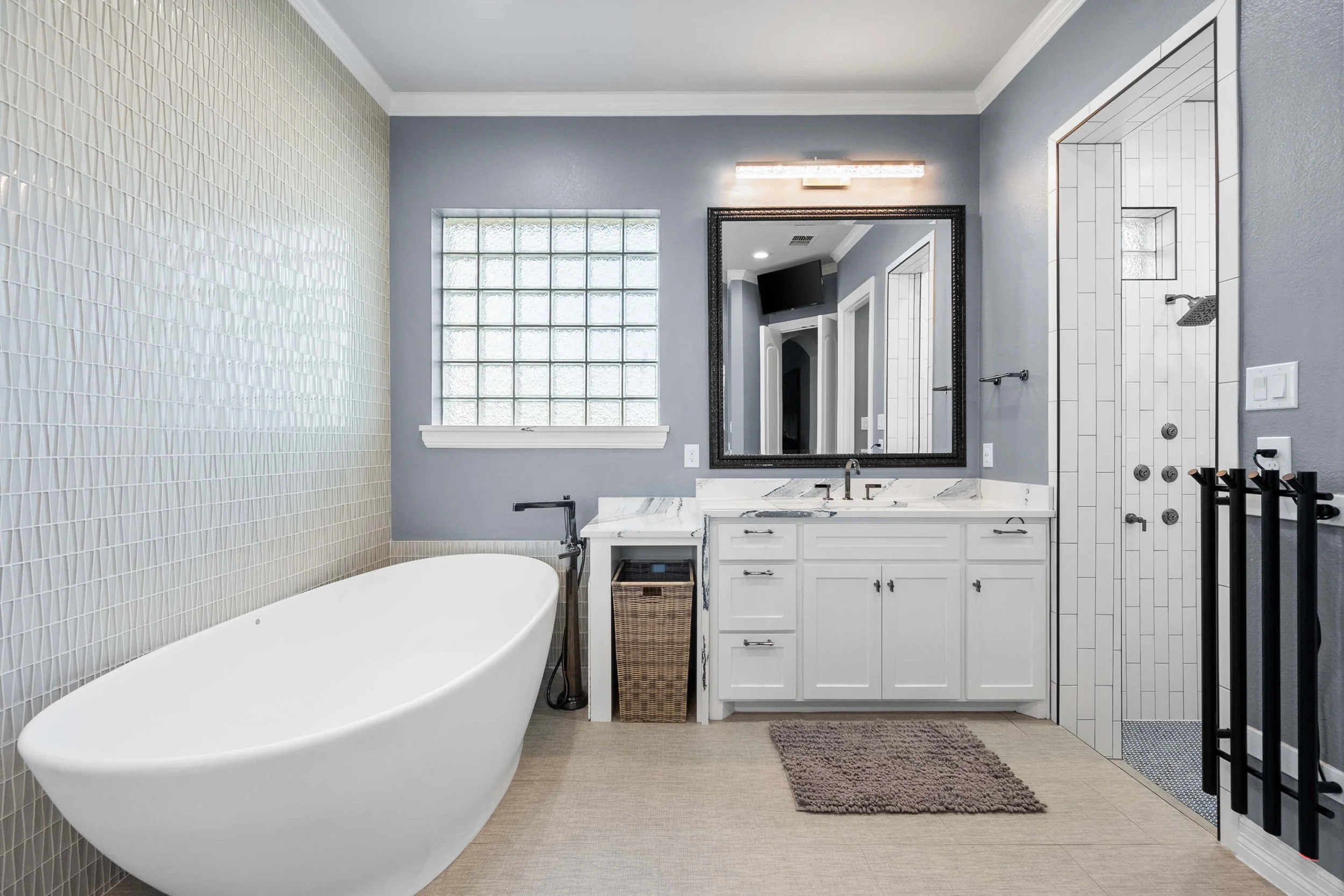 Modern bathroom with a free-standing bathtub, a gray wall, a large glass block window, a white vanity with a marble countertop, a black framed mirror, a shower with white tiles and multiple shower controls, and a brown bath mat.