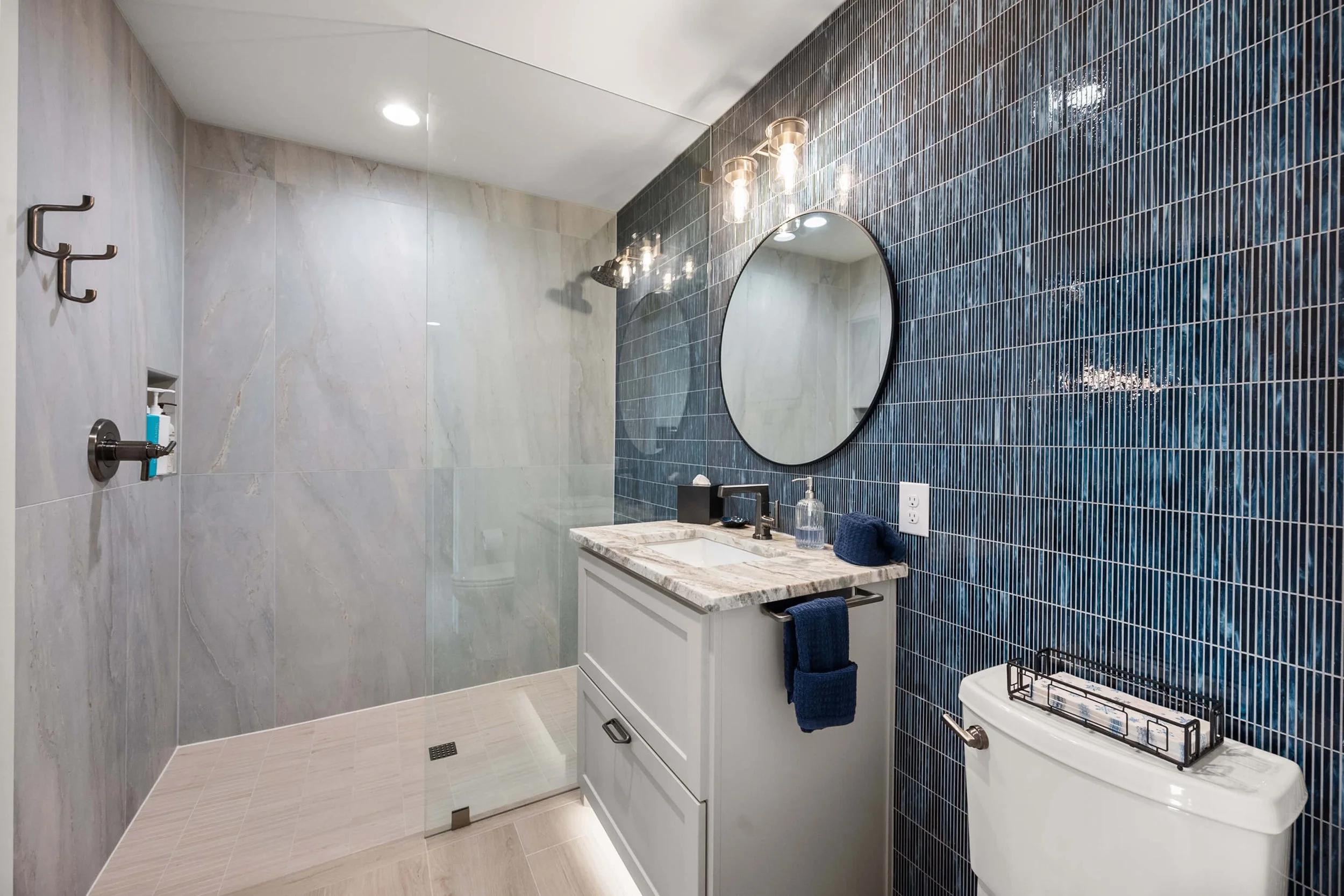 Modern bathroom with a walk-in shower, a vanity with a marble countertop, a round mirror, blue tiles on one wall, and a toilet built by Lakeland Custom Homes.