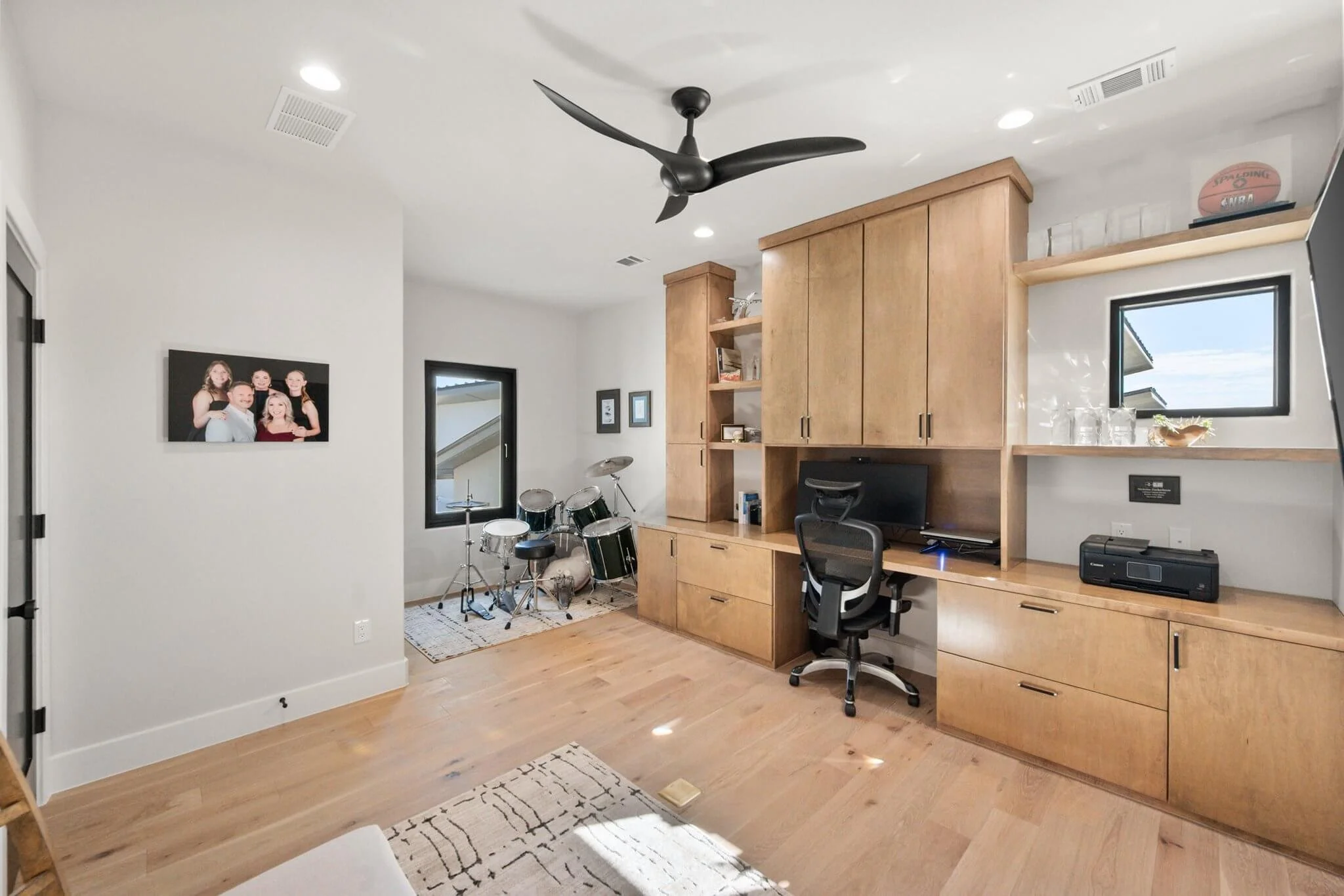Home office with desk, computer, office chair, and wooden storage cabinets, with a small window and a drum set in the corner.