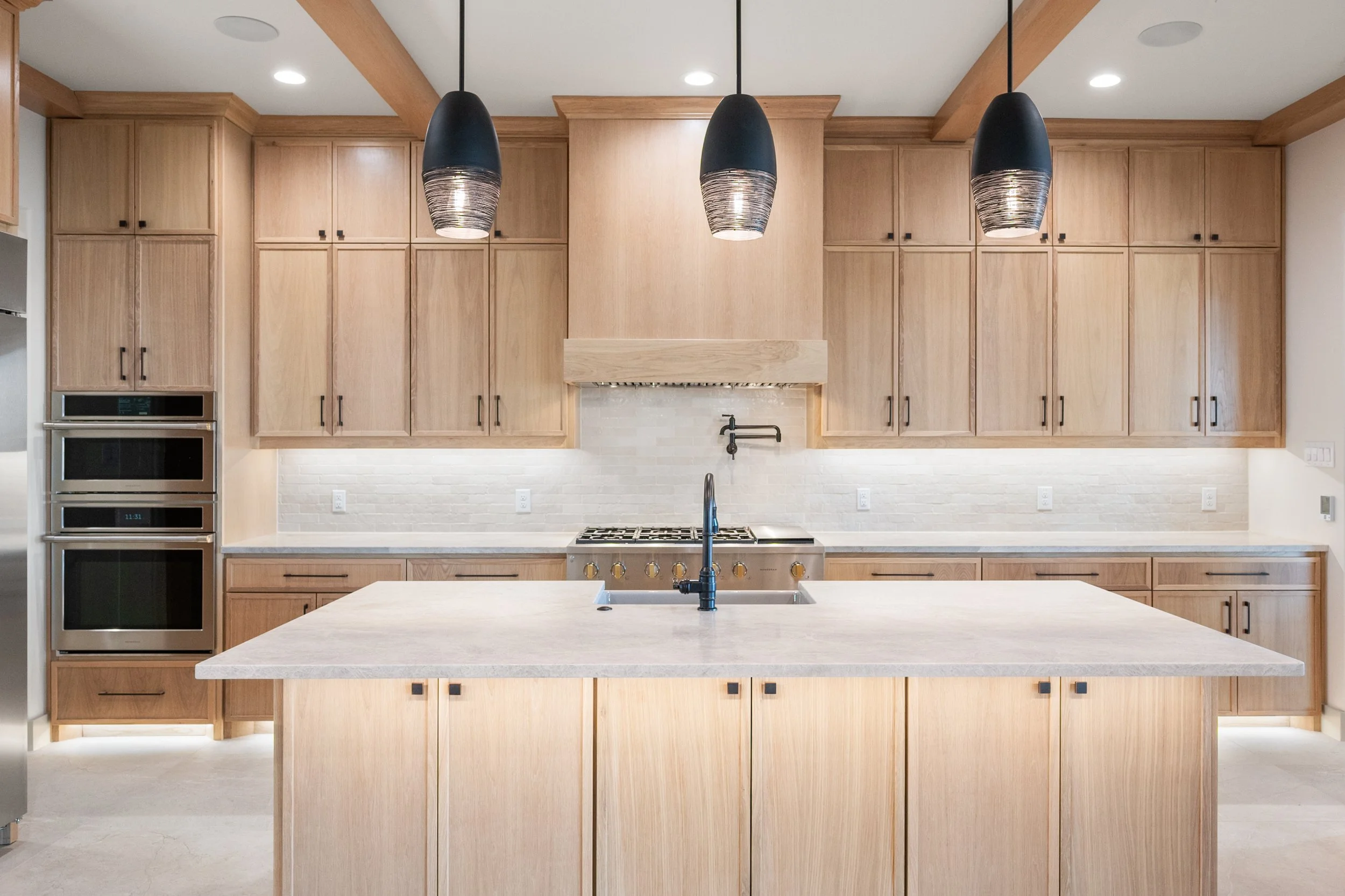 Modern kitchen with light wood cabinets, a large kitchen island with a light-colored countertop, black pendant lights, and built-in stainless steel appliances built by Lakeland Custom Homes.