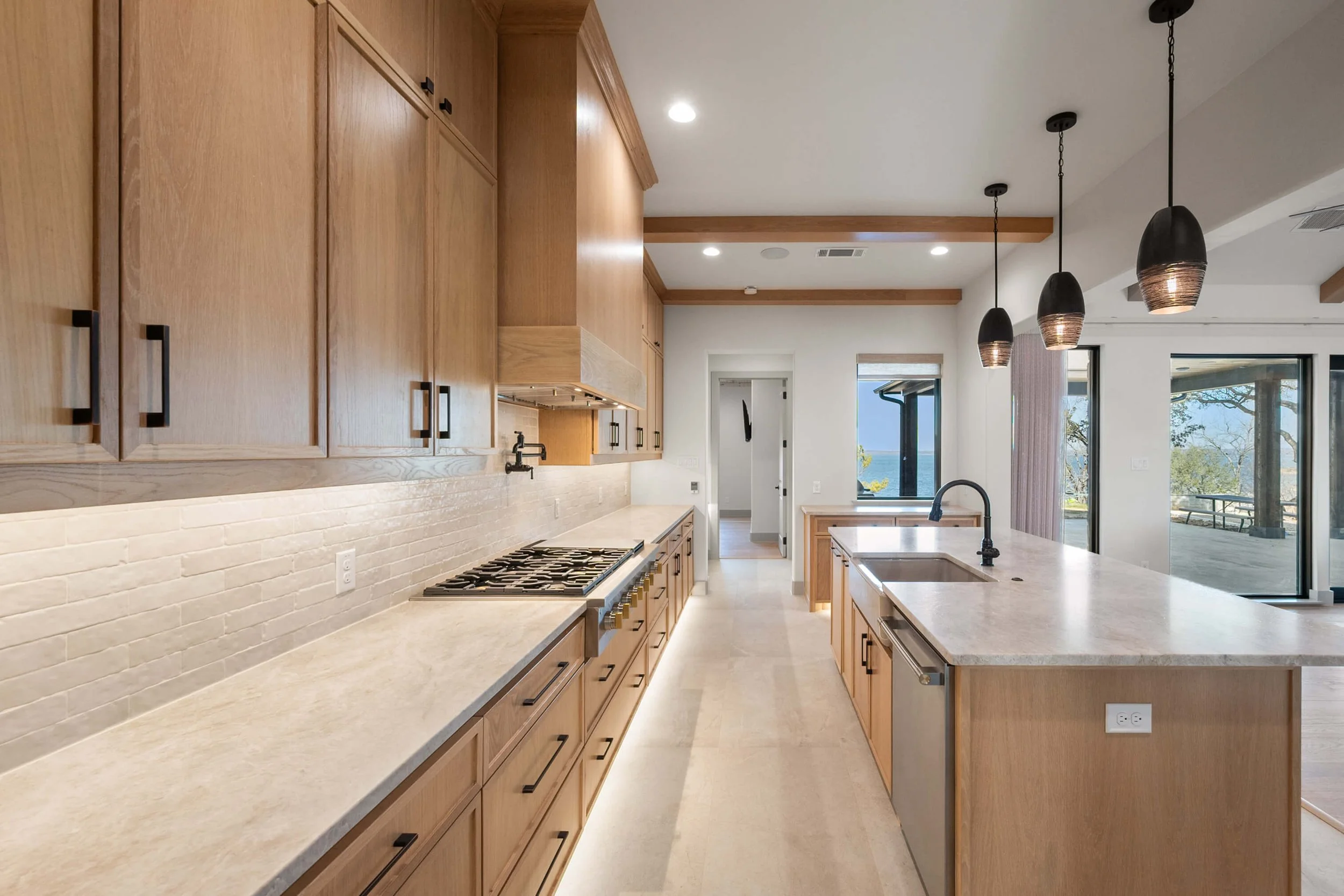 Modern kitchen with wooden cabinets, beige countertops, a gas stove, a sink on an island, black pendant lights, and large sliding glass doors leading to an outdoor deck with scenic views built by Lakeland Custom Homes.