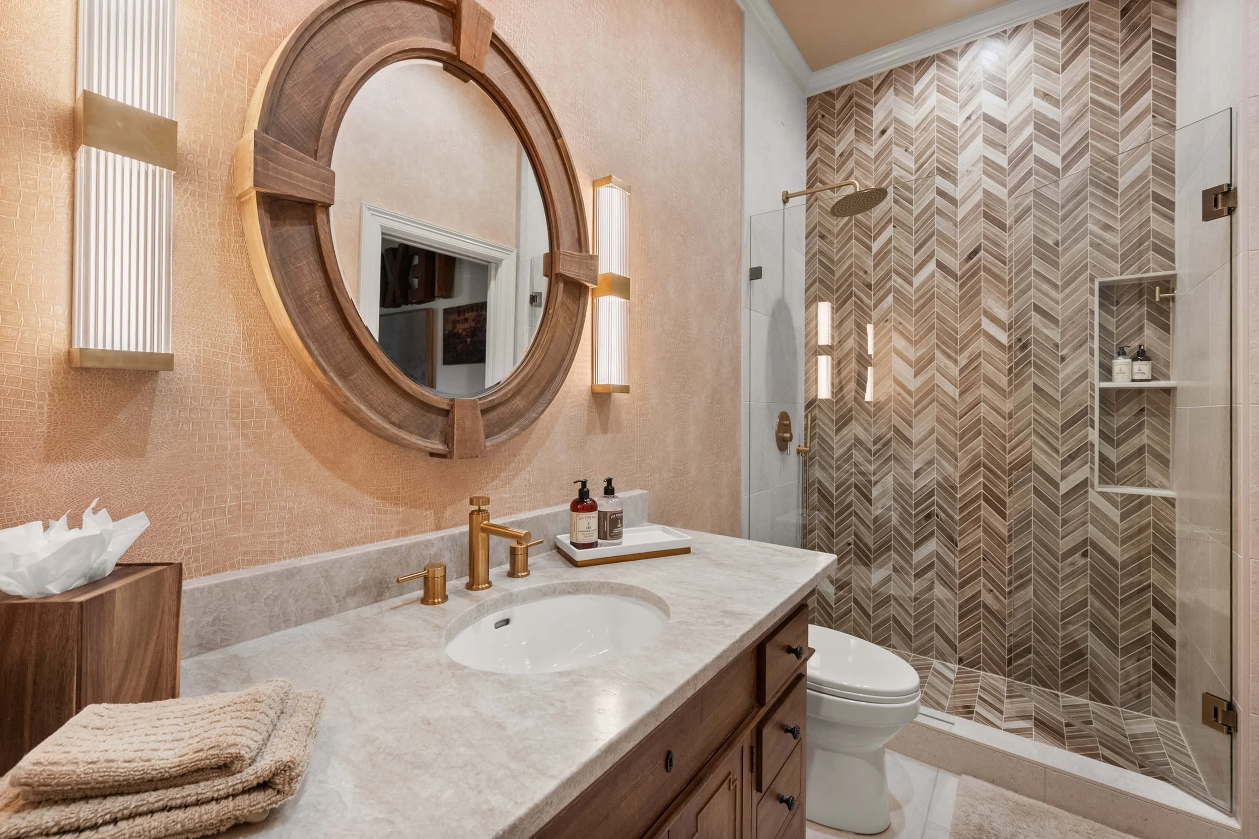 Modern bathroom with a beige marble vanity, a large round wooden-framed mirror, gold fixtures, a toilet, and a walk-in shower with herringbone tiles and built-in shelves.
