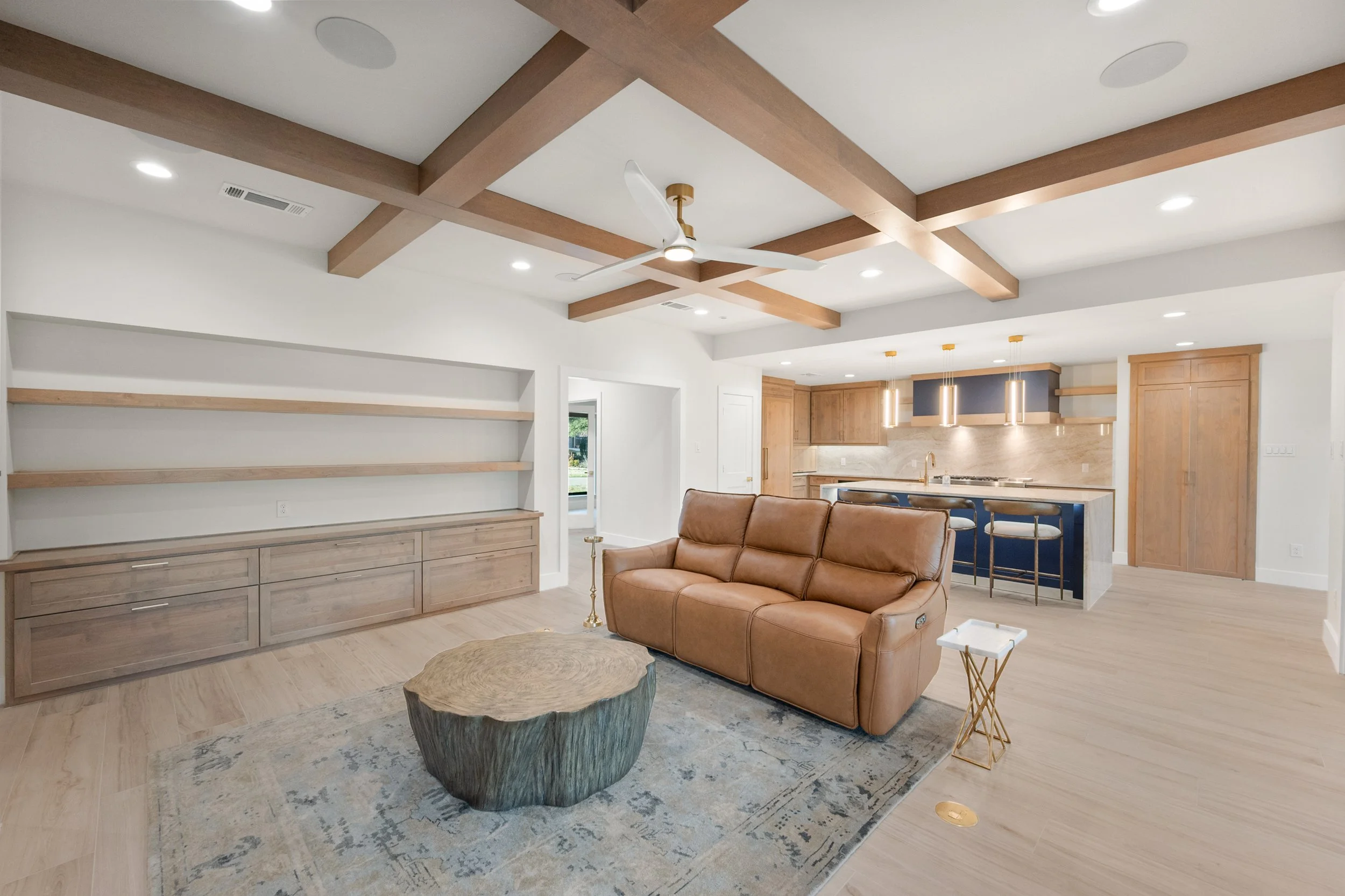 Open-concept living room and kitchen with beige wood accents, a brown leather sofa, a unique wood coffee table, built-in shelves, navy blue and natural wood kitchen cabinets, and pendant lighting.