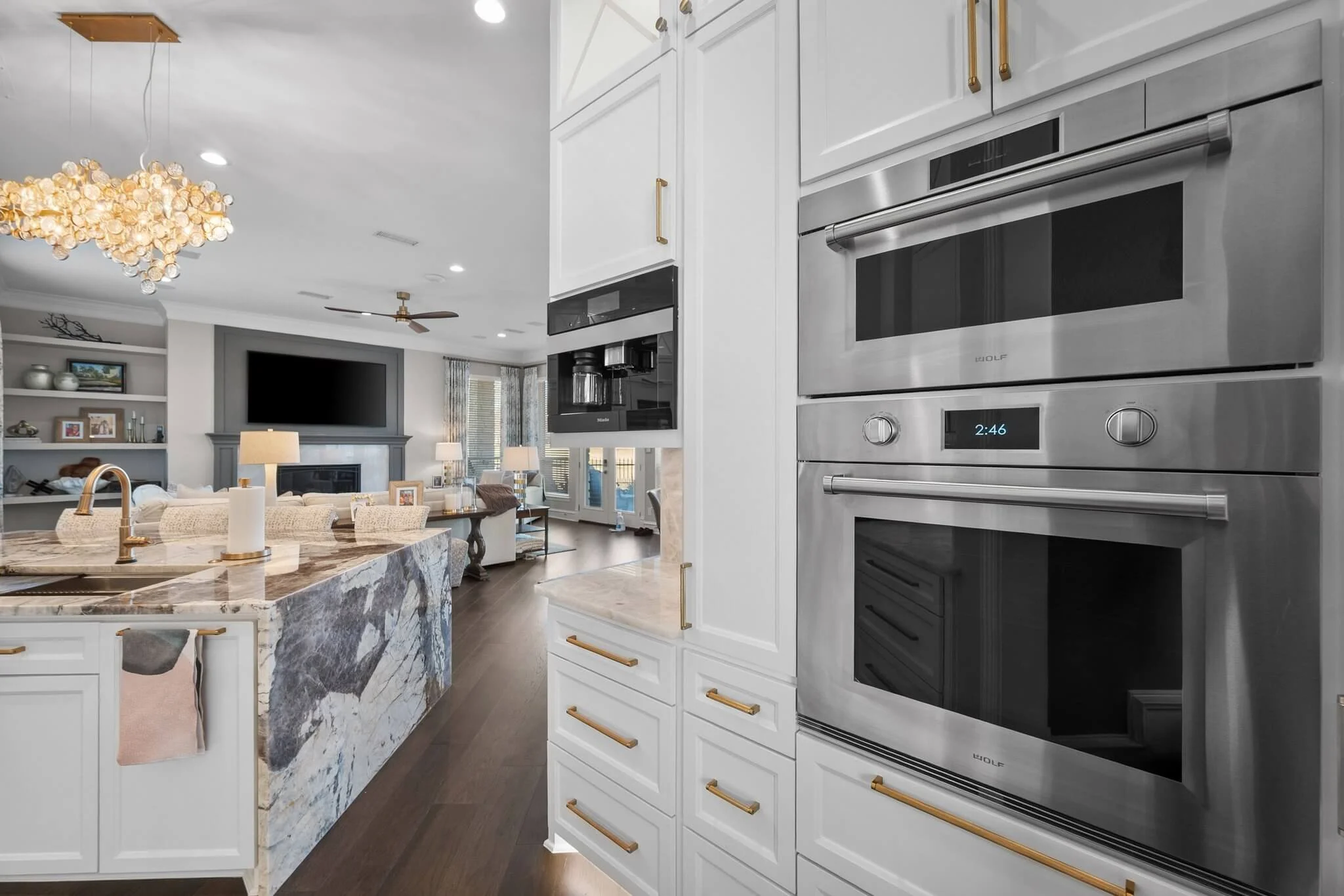 Modern open-concept living space with a kitchen island, white cabinetry with gold handles, stainless steel double oven, coffee machine, dark hardwood floors, a large wall-mounted TV, and a living room area with a fireplace, sofa, and multiple lamps.