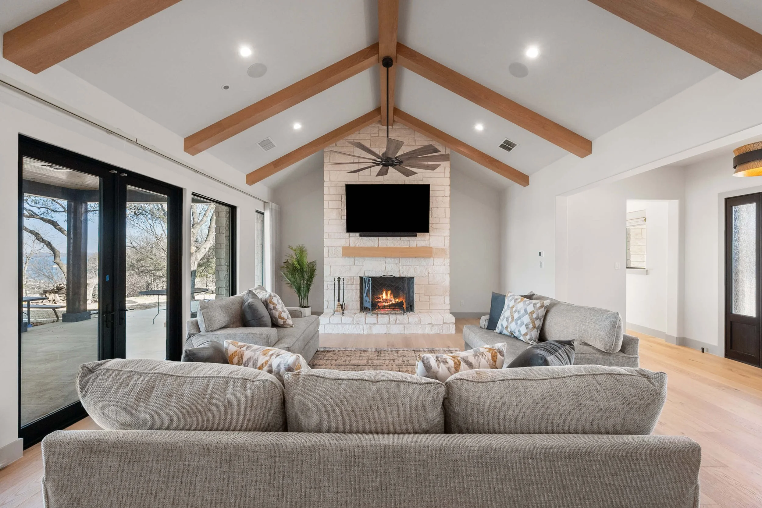 Living room with beige sectional sofa, pillows, glass sliding doors, fireplace, TV, ceiling fan, and wooden beams built by Lakeland Custom Homes..