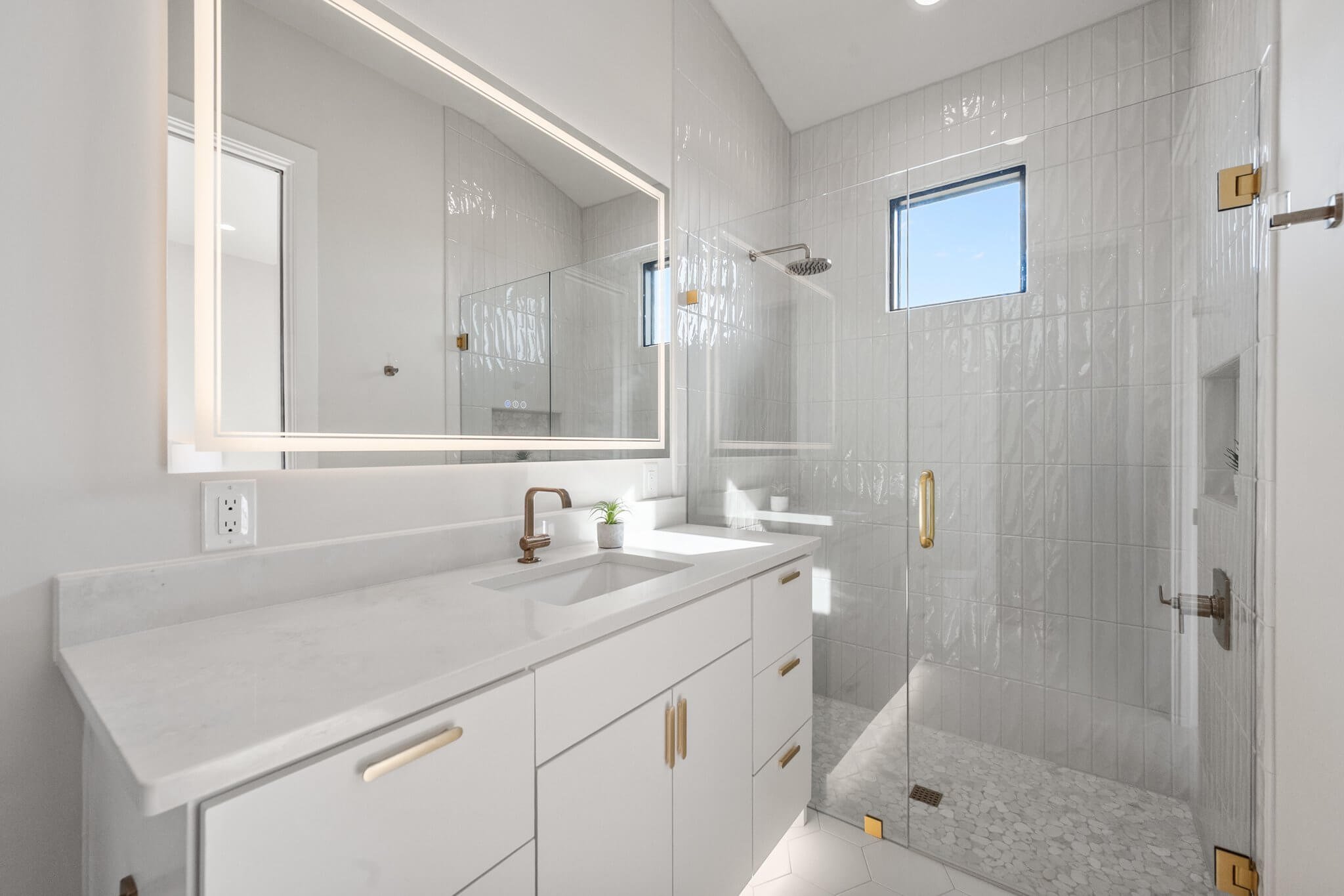 Modern white bathroom with a vanity, large mirror, copper faucet, small plant, and a glass shower with small square tile flooring, a window, and brass accents.